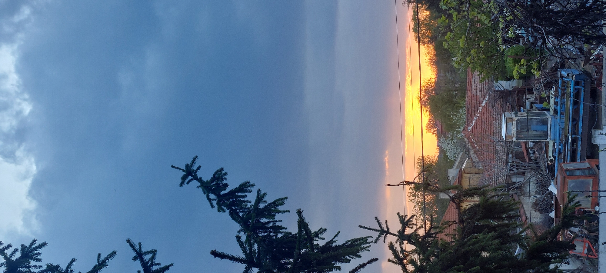 The image captures a sunset scene in Yagodovo, Bulgaria. A clear distinction is visible between the blue, cloudy sky and the horizon, where the sun sets in a bright orange glow. In the foreground, evergreen trees are partially visible. The background reveals a cluttered environment, including buildings with red-tiled roofs, trees, and assorted machinery. Power lines are visible across the sunset.