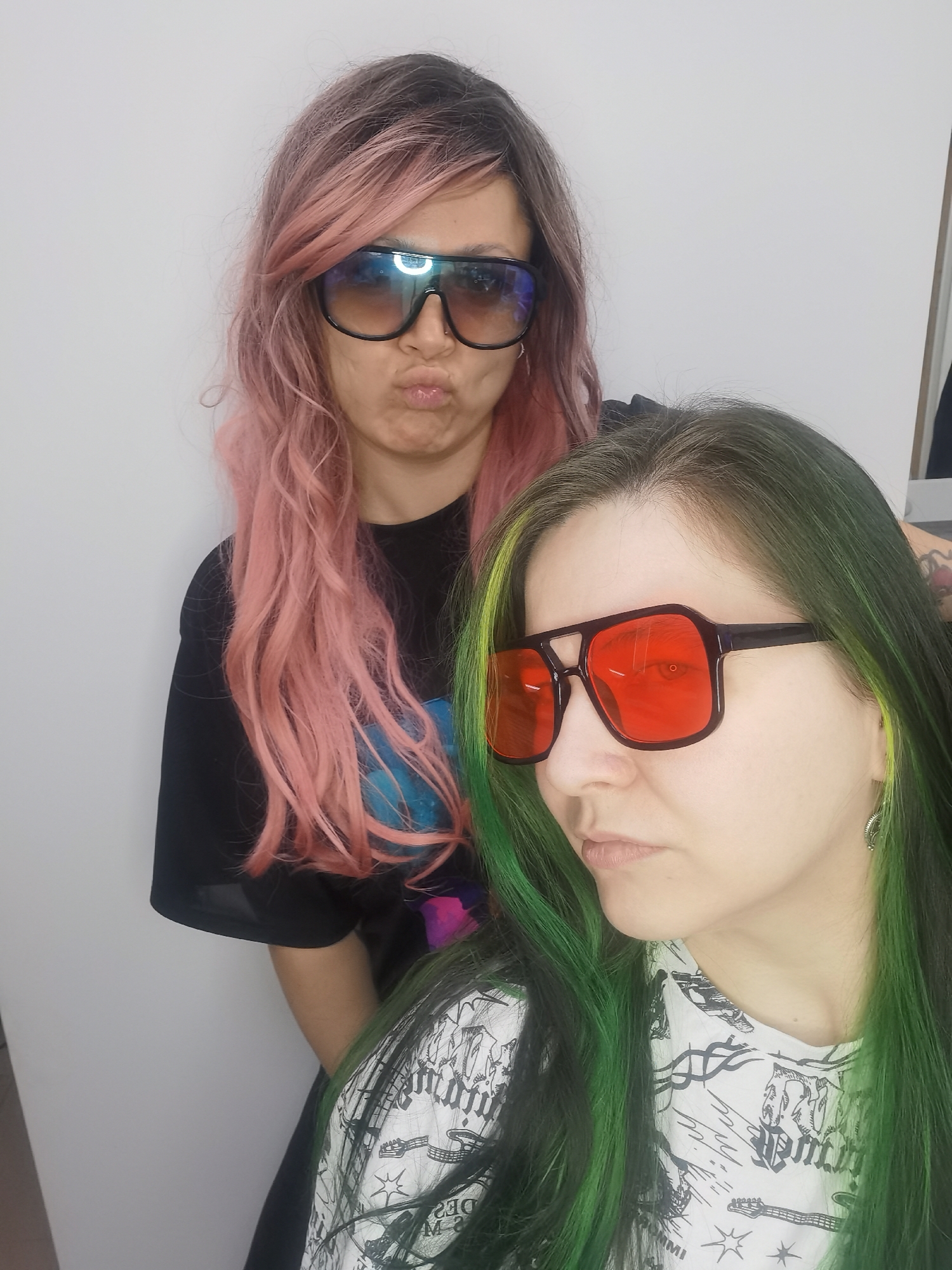 Two women are posing indoors. The woman in the back has long, wavy, pink hair with dark roots, and is wearing dark sunglasses with a thick black frame and tinted lenses. She is pouting her lips. She is wearing a black t-shirt with a graphic print. The woman in front has long, green and black hair with neon green highlights. She is wearing a white t-shirt with a black print. She is wearing dark sunglasses with a thick black frame and red lenses. The background is a plain white wall. The location is the city of Chaikovsky, Russia.