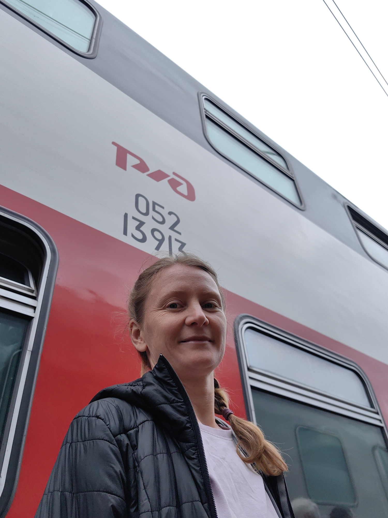 A medium shot captures a woman with blonde hair styled in a braid, standing in front of a Russian Railways (RZD) double-decker passenger train. The train is painted with a red stripe on the lower half and a gray stripe on the upper half, with the RZD logo and the number "052 13913" visible on the side. The woman is wearing a black puffer jacket over a light-colored shirt. The location is Rossosh, Russia, as indicated by the RZD train, which operates in that region.