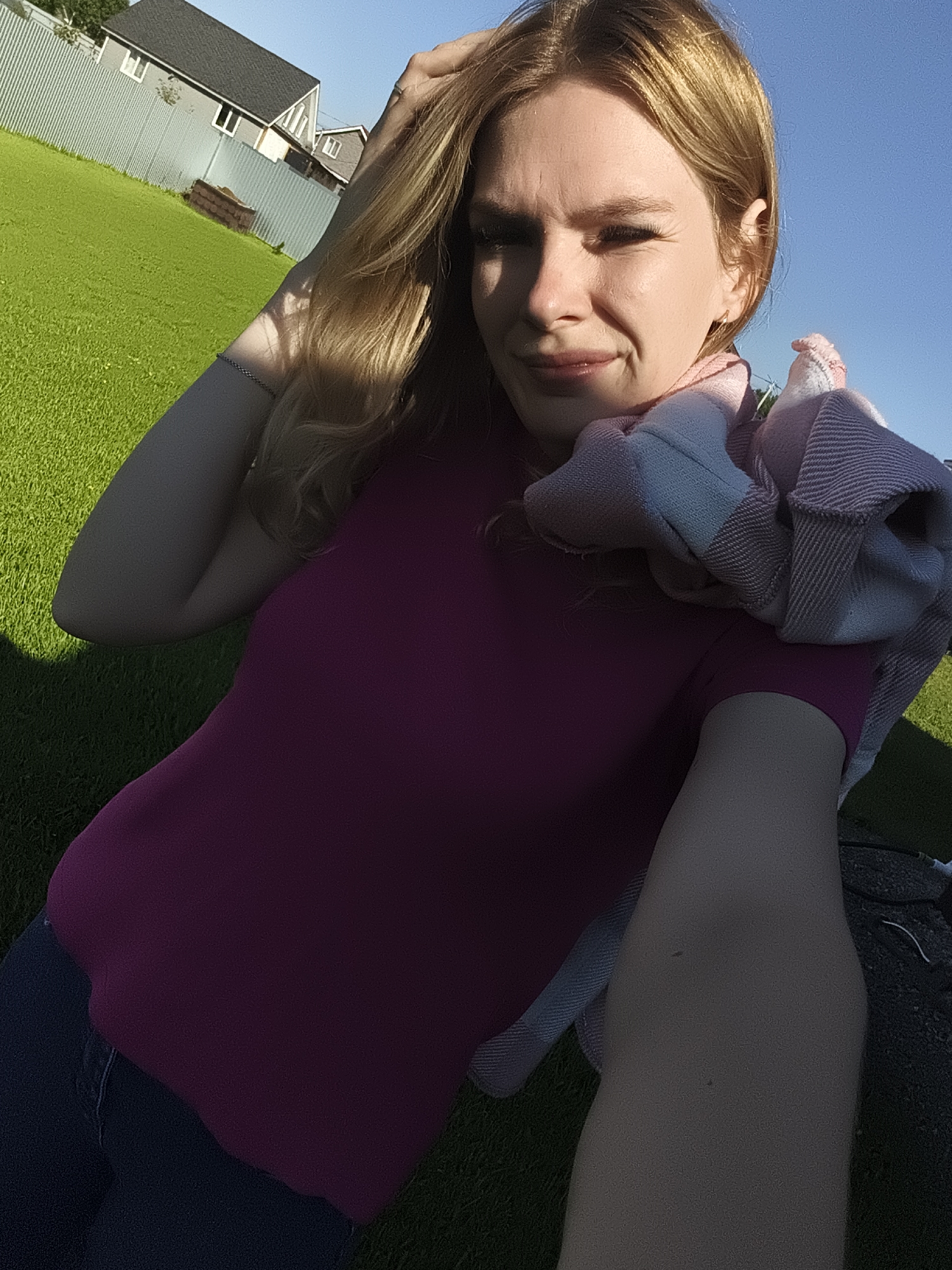 A young Caucasian woman with blonde hair and fair skin is captured in a selfie outdoors, likely in a residential backyard in Русский Вожой, Russia. She is wearing a magenta t-shirt and blue jeans, with a pink and white plaid scarf draped around her neck and shoulders. Her left hand is raised, touching her hair, while her right arm is resting on her leg. The background features a well-maintained green lawn, a grey metal fence, and residential houses with dark roofs, indicative of a suburban or rural setting within the city. The lighting suggests it is late afternoon or early evening, with the sun casting long shadows and illuminating the scene with a warm glow. The sky is clear and blue.