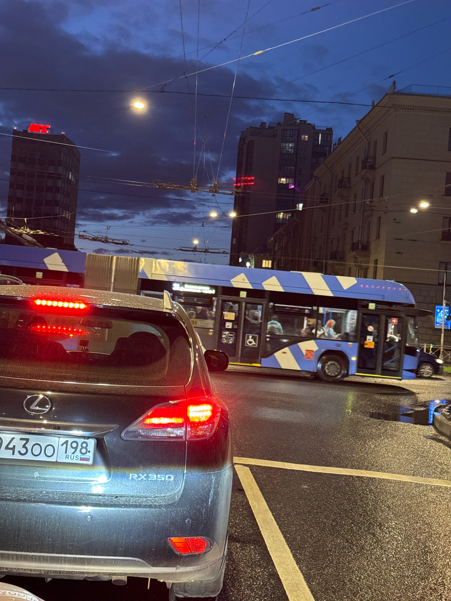 The image depicts a street scene in Saint Petersburg, Russia, at dusk or during twilight. In the foreground, the rear of a dark-colored Lexus RX350 is visible, its red taillights illuminated. The car's license plate reads "94300 198 RUS". Beyond the Lexus, a blue and white striped bus, possibly a trolleybus due to the overhead wires, is stopped in traffic. Several passengers are visible inside the bus. In the background, tall buildings line the street, some with illuminated windows. The sky is dark blue with scattered clouds, and streetlights are on. The road surface appears wet, suggesting recent rain. The overall atmosphere is that of a typical urban evening commute.