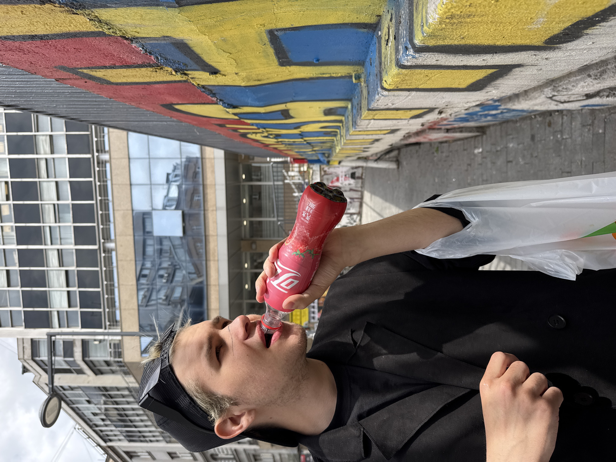A man with light blond hair is drinking from a pink-labeled bottle. He is wearing a black cap with dark straps, a black jacket, and holding a white plastic bag. The backdrop includes buildings with windows and a concrete wall painted with red, yellow, and blue graffiti. The location is the city of Belgrade, Serbia.