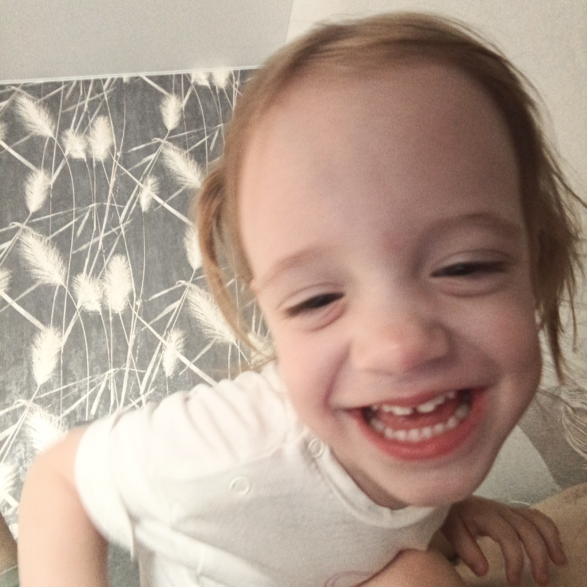 The image is a close-up, front-facing selfie of a young child with blond hair. The child is wearing a white t-shirt with snap closures. The background consists of light-colored wallpaper with a repeating pattern of white grass-like stalks against a gray base. The child's face is large in the frame, with their mouth open in a wide smile, revealing their teeth. The lighting appears to be natural, coming from the front. The location in Марковский, Russia is not discernible from the image itself.