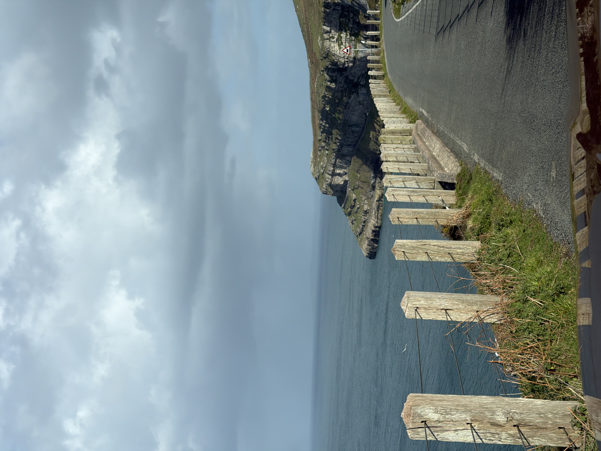 The image captures a coastal scene in Llandudno, United Kingdom. A paved road runs along the edge of a cliff. Adjacent to the road is a low, wooden fence constructed with horizontal planks supported by posts. Below the fence, a steep cliff drops to the sea. The sea is visible, extending towards the horizon under a cloudy sky. A road sign is noticeable near the top of the cliff. The photograph is oriented vertically.