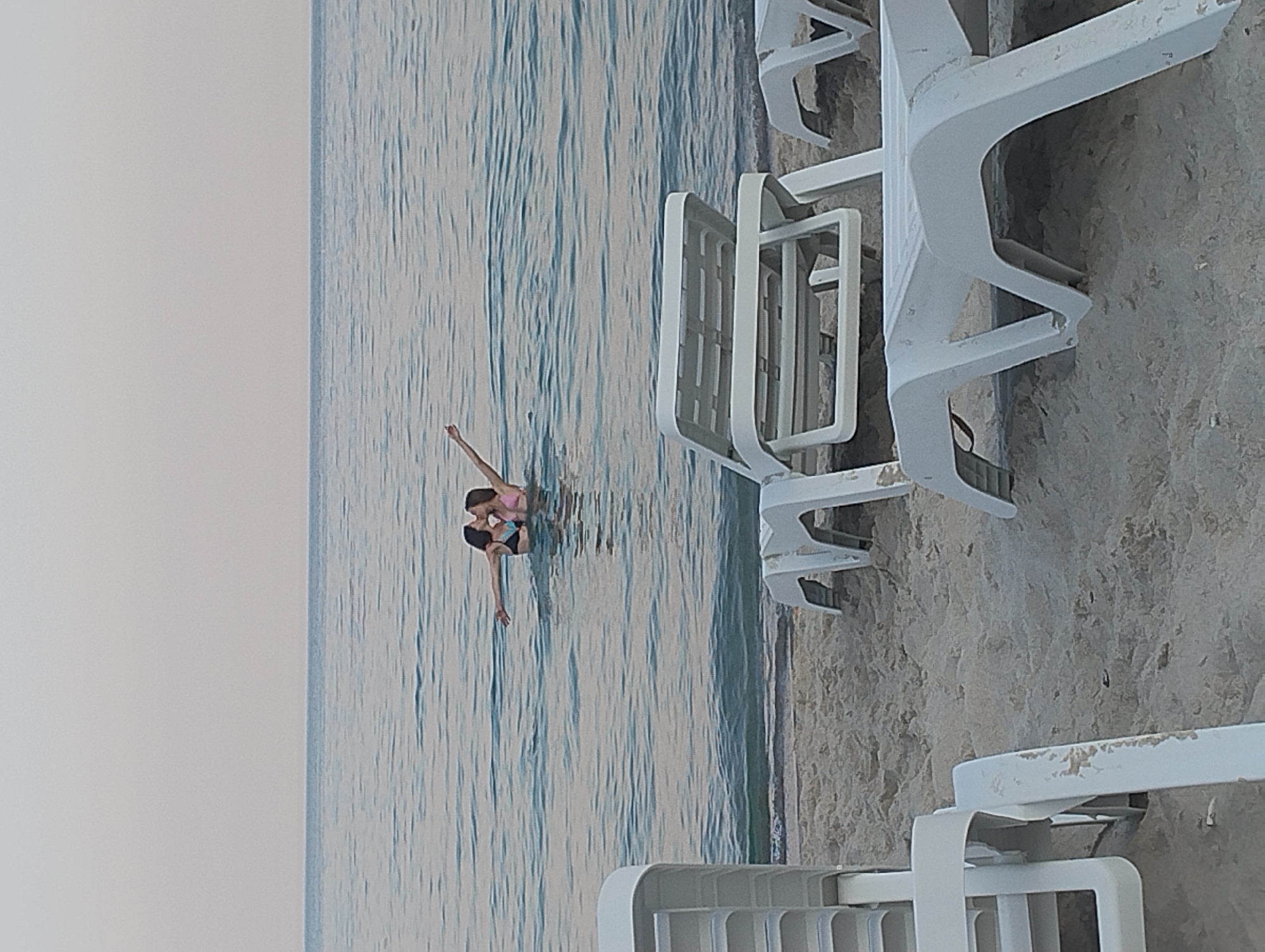 In Odesa, Ukraine, on a sandy beach with the calm sea stretching to the horizon under a pale sky, two women are in the water. They are dressed in bikinis and appear to be embracing or kissing. In the foreground, several white plastic lounge chairs are visible, some stacked and others arranged on the sand, suggesting a resort or public beach setting. The water is a muted blue-gray, reflecting the overcast sky.