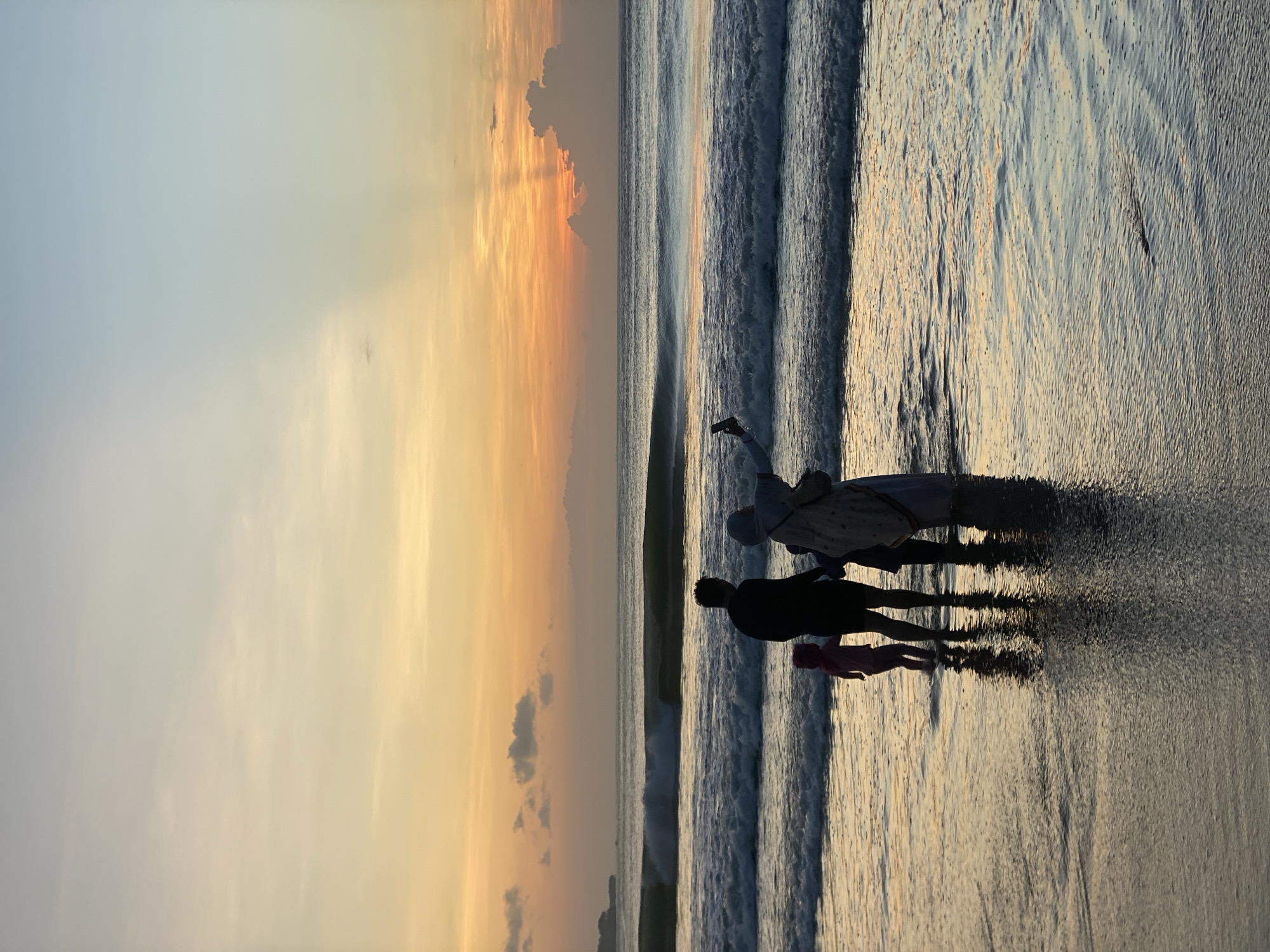 The image shows a beach scene at sunset. The location is likely in or near Kuta Selatan, Indonesia, known for its beaches. There are three figures in silhouette standing on the wet sand near the water's edge. One appears to be an adult wearing a patterned garment and possibly a head covering, holding an object aloft. A second, shorter figure stands beside them, and a small child in pink stands next to them. The sky is a gradient of blue to orange, indicating sunset, with rays of light extending downward. The ocean has small waves coming in to the shore, reflecting the sunset colors.