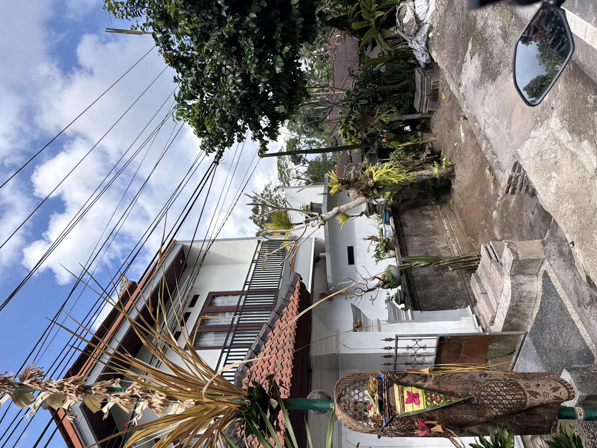 The image depicts a residential area in Susut, Indonesia, with a focus on a dwelling featuring a white facade and a red-tiled roof. A prominent element in the foreground is a decorative pole draped with brown fabric, adorned with a basket containing offerings. Adjacent to the house, a balcony with a black railing is visible, and above, a network of black utility wires crisscrosses the sky. The scene is filled with lush greenery, including trees and various potted plants. The ground is a mix of dirt, gravel, and concrete pathways. A motorcycle mirror is visible in the lower right corner, reflecting trees and a person. The overall impression is one of a typical tropical environment with elements of local culture evident in the decorative offerings.