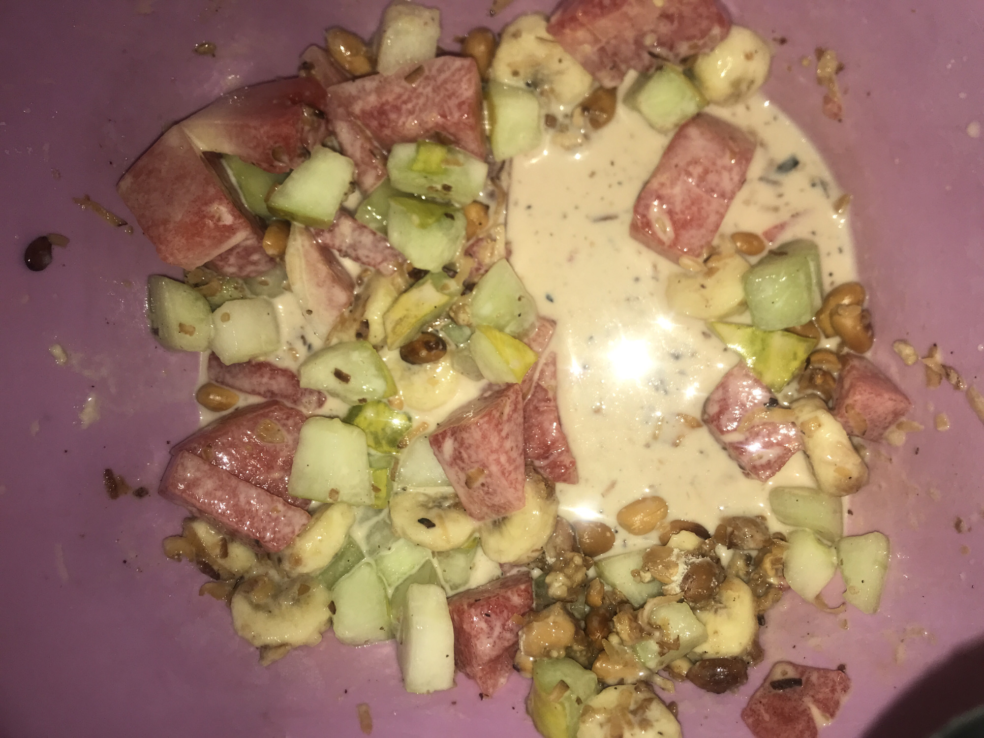 The image presents a close-up, overhead view of a fruit and nut mixture served in a light purple bowl. The scene focuses entirely on the food, with no people visible.
The dish consists of an assortment of diced fruits and various nuts, partially immersed in a creamy, light beige liquid. Prominent fruit components include chunky, reddish-pink cubes of watermelon, slices of yellowish-white banana, and light green, cubed pieces resembling cucumber or a similar melon. Scattered among the fruit are numerous nuts, notably oval-shaped, light brown peanuts, along with smaller, irregularly shaped brownish pieces that could be granola, cereal, or other mixed nuts. The creamy liquid, possibly yogurt or a dairy-based dressing, has dark speckles dispersed throughout, suggesting spices, seeds, or finely ground ingredients.
The activity depicted is the presentation of a prepared fruit salad or dessert, ready for consumption. The items appear thoroughly mixed, with the liquid coating most of the solid ingredients. The purple bowl serves as the sole contextual element for the setting, offering no specific location cues beyond the provided information that it is Rafin Par, Nigeria. The lighting in the image suggests an indoor environment, possibly under artificial light, as indicated by reflections on the liquid surface. There is no visible text in the media file.