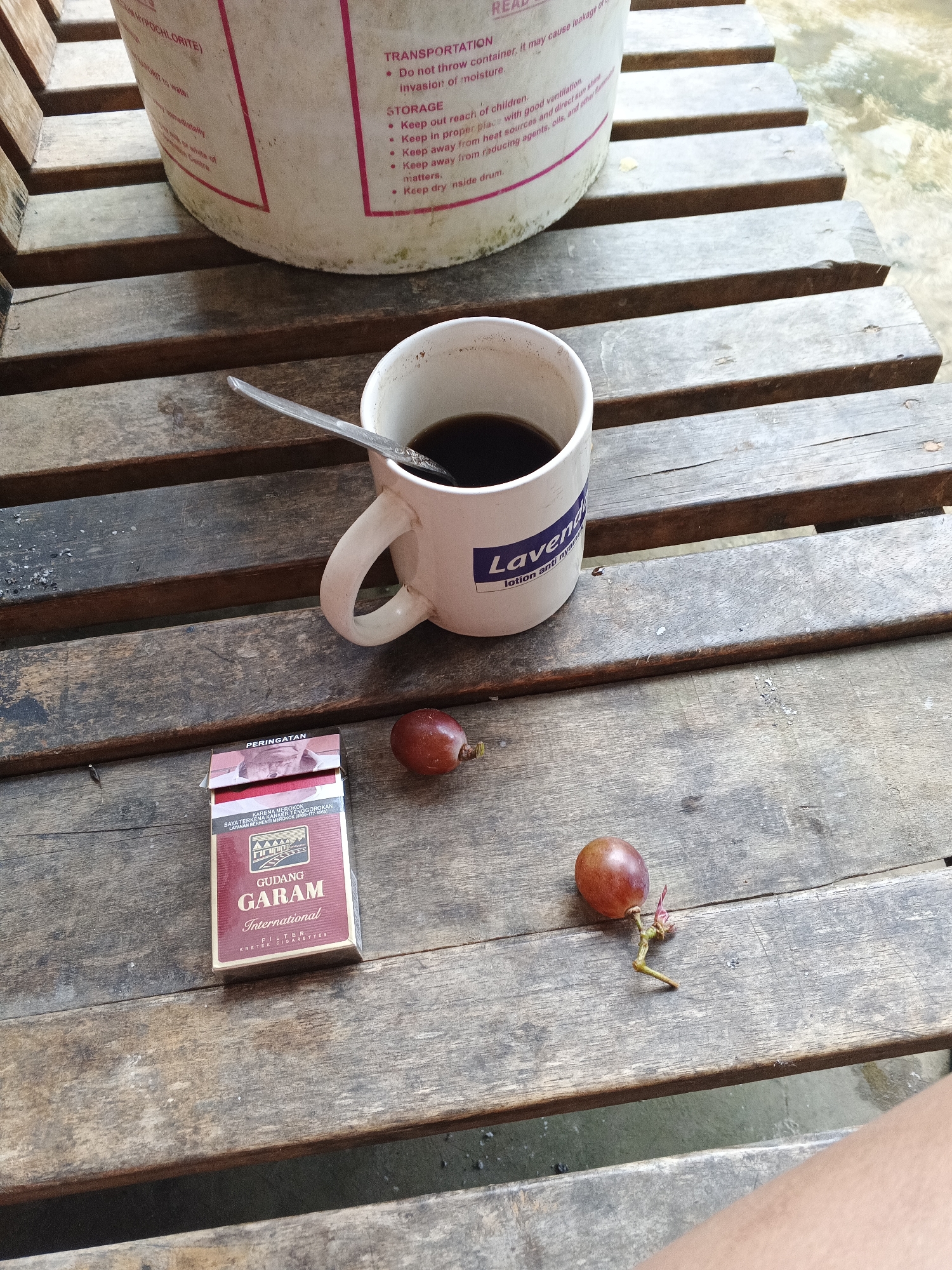 A close-up, high-angle shot captures a rustic wooden table with a mug of black coffee, a pack of Gudang Garam cigarettes, and two red grapes. The coffee mug, white with a blue logo that reads "Lavend...", sits on the table, a silver spoon resting inside. To the left of the mug is a red and gold pack of Gudang Garam International kretek cigarettes, open to reveal its contents. Two red grapes, one slightly detached from its stem, are placed on the table between the cigarettes and the mug. In the background, the edge of a large white plastic bucket with red lettering is visible, and beyond that, a body of murky water. The wooden planks of the table are weathered and spaced, revealing glimpses of the ground beneath. This scene is likely set outdoors, possibly in a rural or semi-rural setting within Warungkiara, Indonesia, given the visible branding on the cigarette pack.