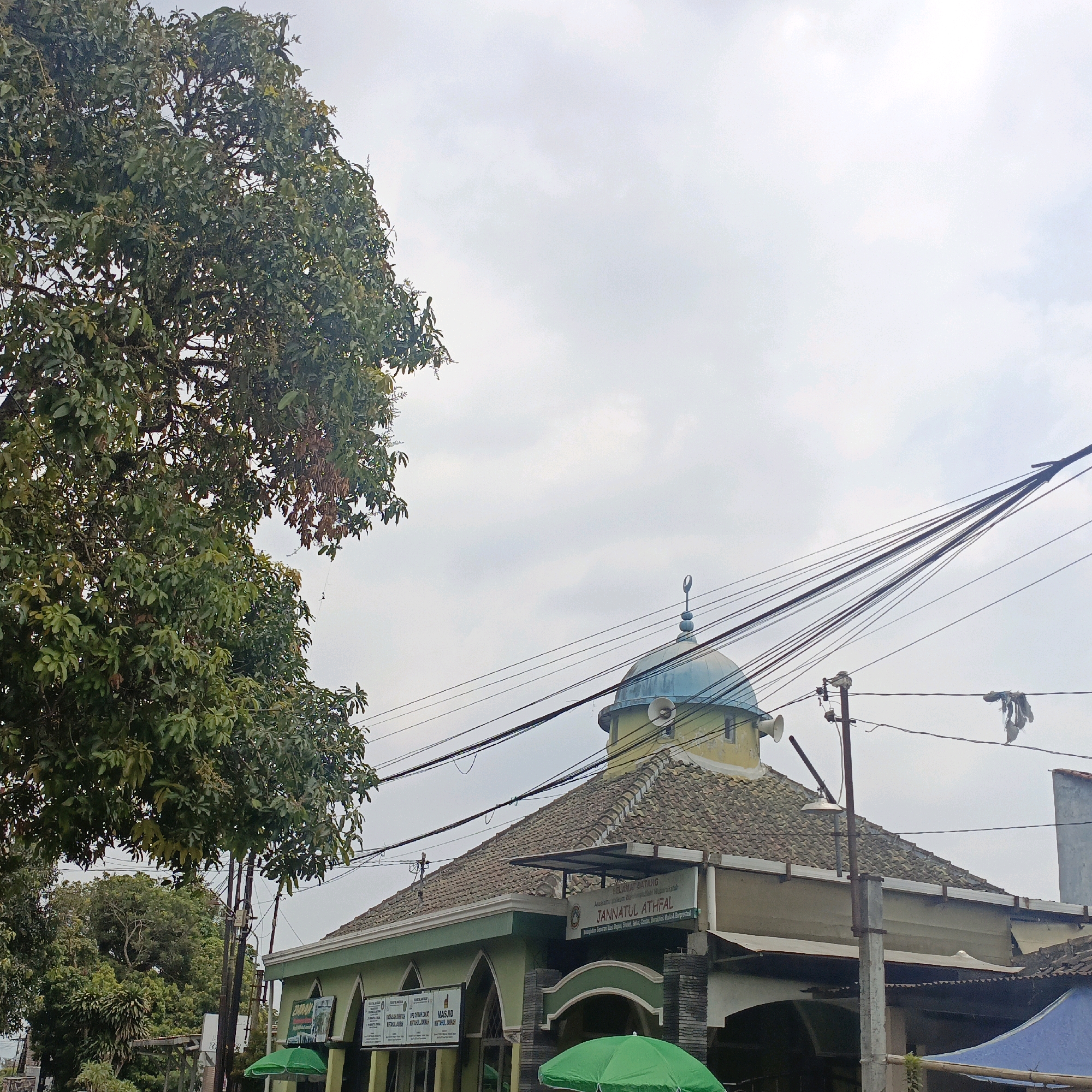 The image shows a mosque with a green and yellow facade and a blue dome, located in Salatiga, Indonesia. The mosque is situated on a street with overhead electrical wires and a large tree with green foliage in the foreground. There are signs on the mosque, one of which reads "SELAMAT DATANG" and "JANNATUL ATHFAL". Two green umbrellas are visible in front of the mosque. The sky is overcast.