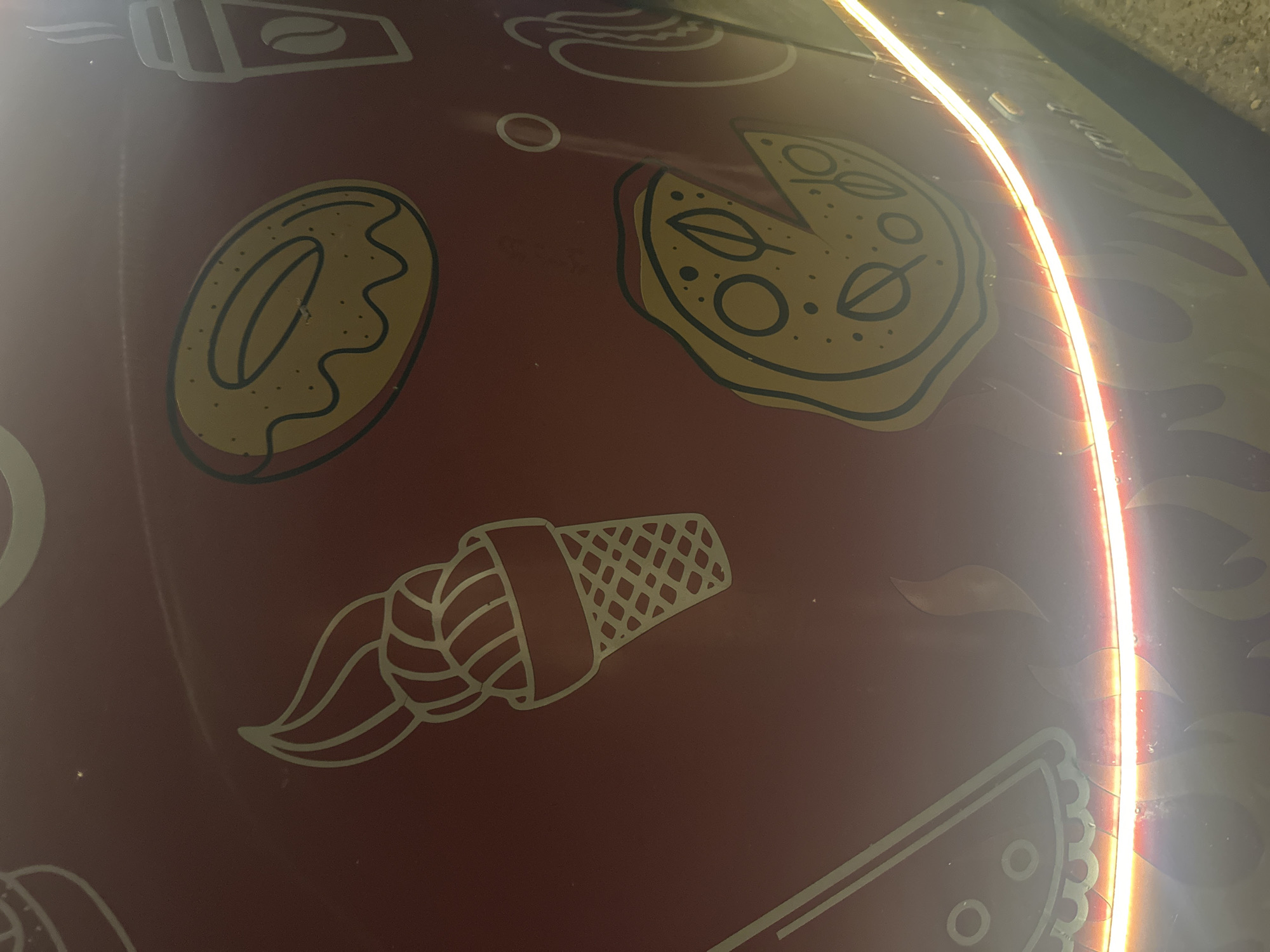 The image shows a close-up of a red surface with illustrations of food items, specifically a pizza slice, an ice cream cone, a donut, and a coffee cup. These drawings appear to be stylized and outlined in a lighter color, possibly gold or yellow. The surface is curved and reflects a bright orange light, suggesting it might be part of a vehicle or a kiosk. The background is dark and out of focus. The context of Tomsk, Russia, is not evident from the image itself. The image is an indoor or outdoor shot, given the lighting conditions and the textured ground visible at the bottom.