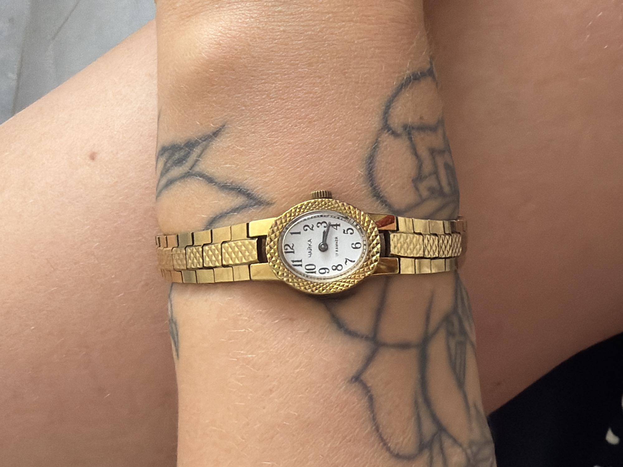 A close-up, eye-level shot shows a gold-colored wristwatch with an oval face on a person's wrist. The watch has a white face with black Arabic numerals and gold hands. The brand name "ЧАЙКА" is visible at the top of the watch face, and "17 KAMNER" is visible at the bottom. The watch band is made of interlocking gold-colored links with a textured pattern. The person's arm, which is tanned and has visible body hair, is adorned with black tattoos, including a floral design and abstract lines. The background is out of focus and appears to be a neutral surface, possibly a wall or floor. The image was taken on a sunny day, as indicated by the lighting. The watch is positioned centrally on the wrist, suggesting it is being worn. The image does not provide any specific information about the city of Sarapul, Russia.