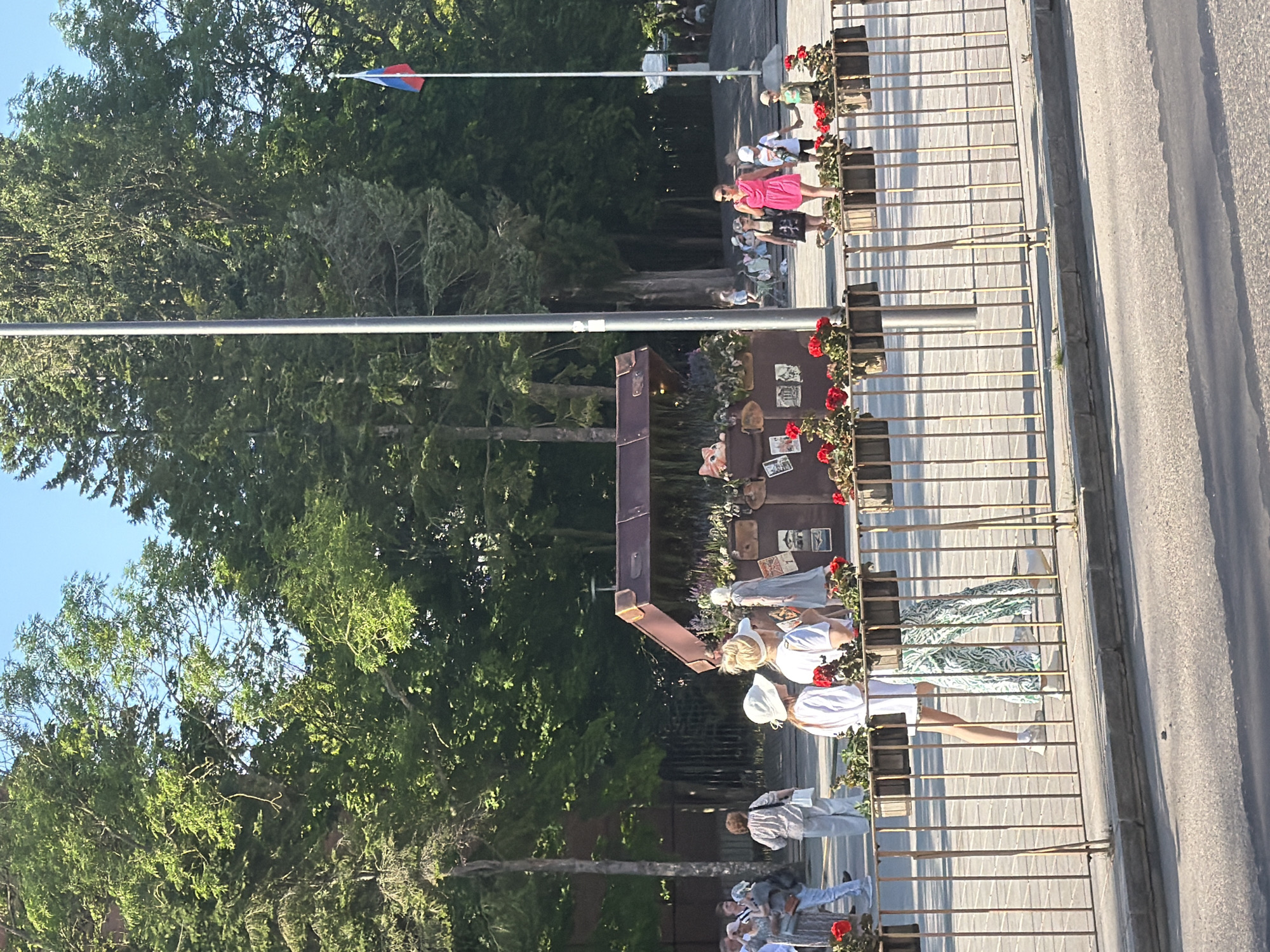In the city of Zelenogradsk, Russia, a group of people are gathered in an outdoor public space on a clear, sunny day. In the foreground, a metal railing with vertical bars separates the viewing area from a paved surface. Behind the railing, a decorative display resembling an open, antique trunk is set up. The trunk is adorned with various items, including plants, flowers (predominantly red), and framed pictures. Two women are prominently visible near this display, one wearing a white hat and white top with patterned green pants, and the other in a white top and white hat.
In the background, more people are dispersed across a paved plaza, with a large number of mature trees providing shade. A flagpole with the Russian flag is visible on the right side of the image. The overall atmosphere appears to be one of leisure and public gathering.