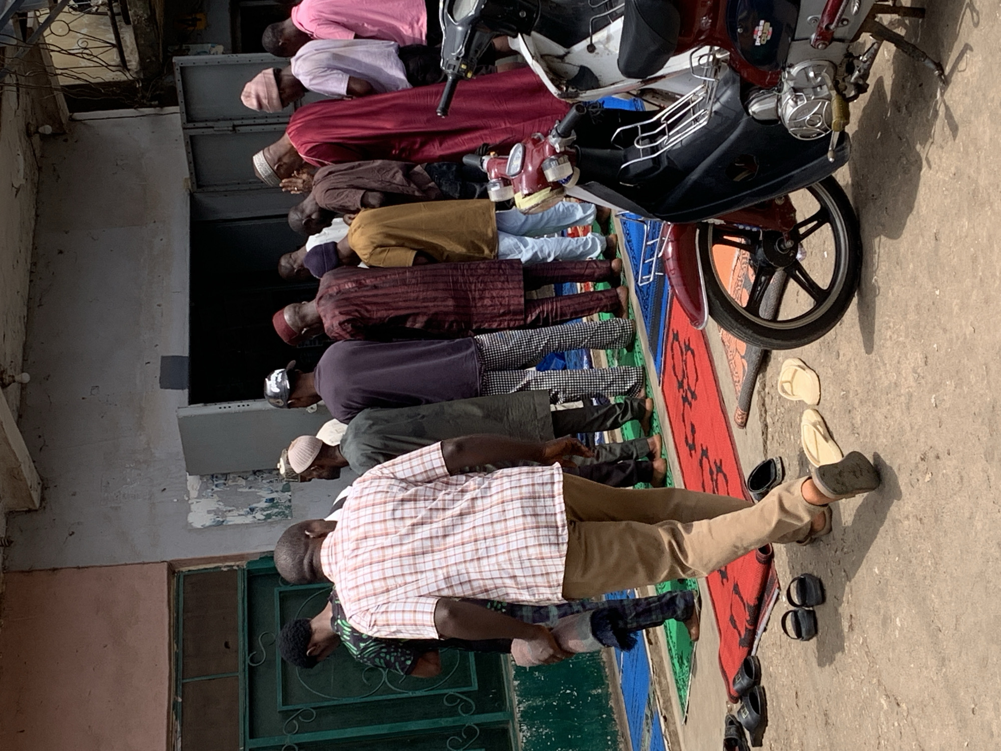 In Jos, Nigeria, a line of at least ten men, many wearing head coverings, are in prayer, bent over in unison, on a multi-colored rug laid out on a concrete surface outdoors, beside a building with a green-framed window and peeling paint. A motorbike is parked nearby. Various pairs of shoes and sandals sit to the side of the rug. The men wear different clothing including pink, red, brown, yellow, striped, and plaid shirts.