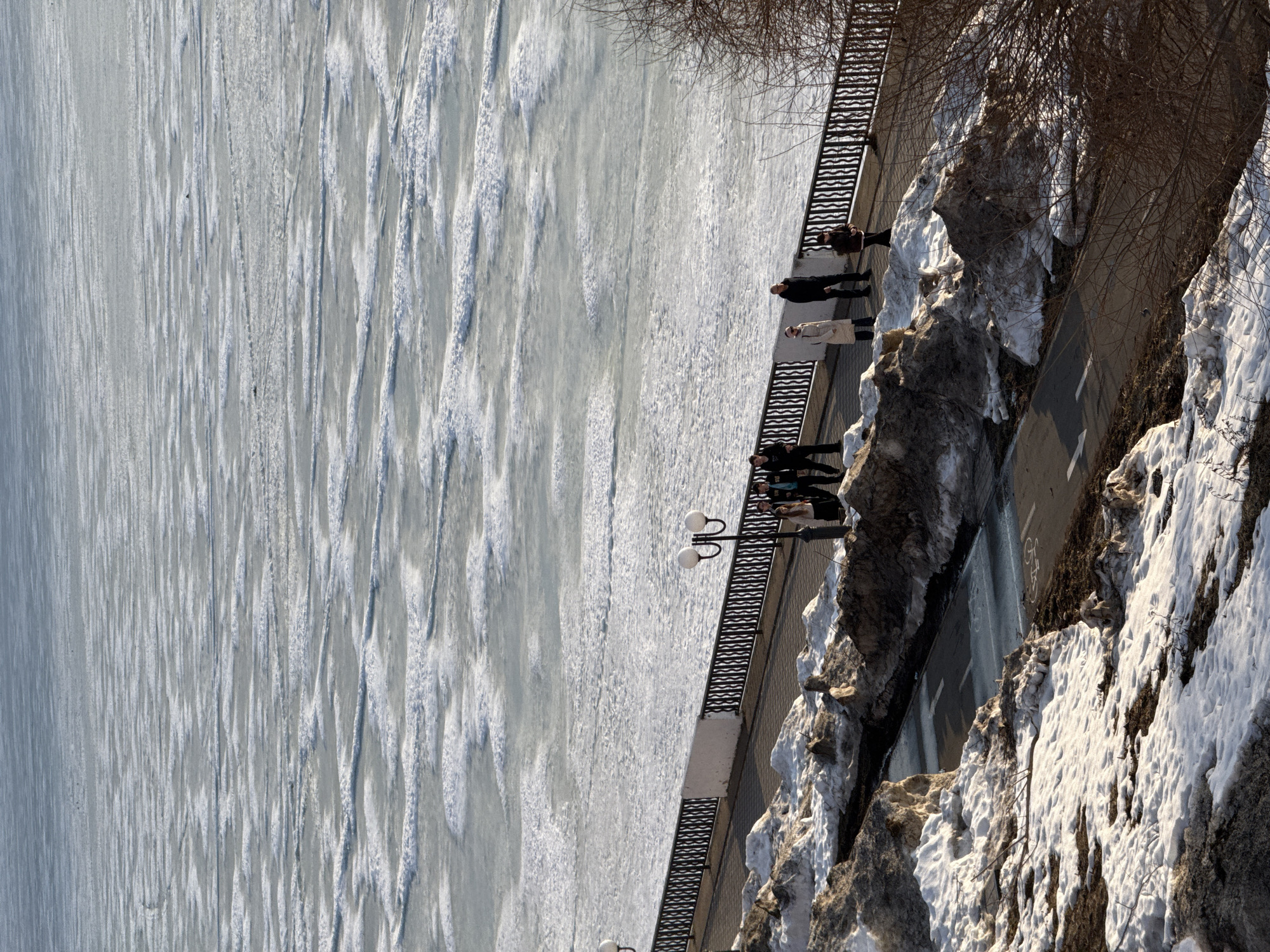 A view of the ice-covered Votkinsk reservoir and the city embankment, Votkinsk, Russia. The ice is light gray and white with long, thin ridges. Several people are standing on the embankment, looking out over the ice. The embankment is lined with a black metal railing and occasional street lights. Snow-covered rocks and bare trees line the side of the embankment. A road can be seen behind the embankment.