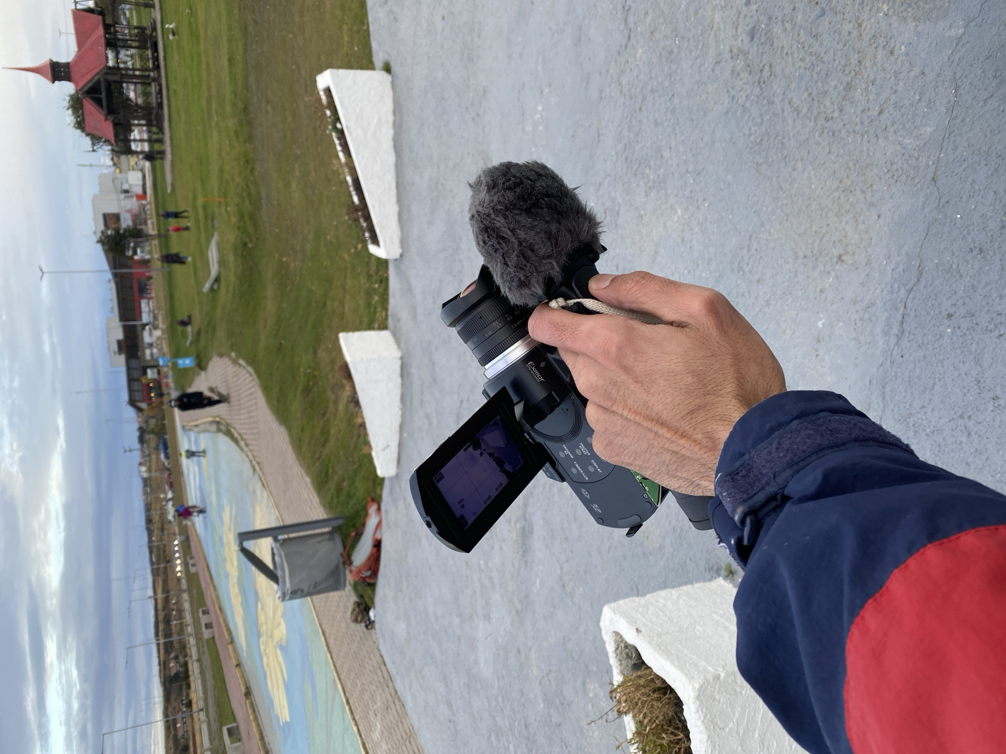 A first-person perspective photograph shows a person holding a video camera outdoors. The camera has a microphone attached with a fuzzy windscreen. The camera screen is flipped open, displaying settings. The person's arm and jacket sleeve, colored red and blue, are visible in the foreground.
The background features a park-like setting with a green lawn, trees, and a paved walkway. A wooden structure with a red roof is visible in the upper left. In the distance, other buildings and streetlights can be seen.
The location is likely Ushuaia, Argentina. The park's painted walkway and distinctive red-roofed structure are landmarks in the city. People are walking in the background.