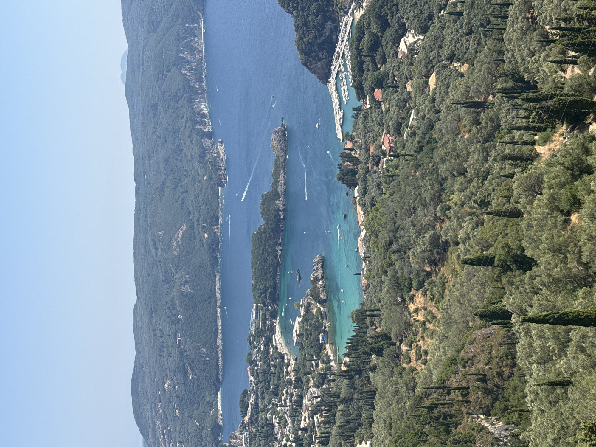 The image captures a sweeping panoramic view of a coastal bay, likely near Λάκωνες, Greece. The foreground is dominated by a lush, green hillside densely covered with olive trees and interspersed with tall, slender cypress trees. Scattered amongst the foliage, red-tiled roofs of houses are visible, suggesting a residential area or a small village nestled within the landscape.
The midground transitions to the vibrant turquoise waters of the bay, which are dotted with numerous small boats and yachts. A small harbor with several docked boats is visible on the right side of the bay. Along the coastline, buildings and structures are built close to the water's edge, with some appearing to be hotels or resorts. Rocky outcrops and small islands emerge from the water, adding to the picturesque scenery.
In the background, rolling, tree-covered hills rise up, forming a dramatic backdrop against the clear blue sky. The distant mountains are hazy, suggesting a warm, sunny day. The overall impression is one of a tranquil, scenic Mediterranean landscape. There are no people clearly visible in the image.