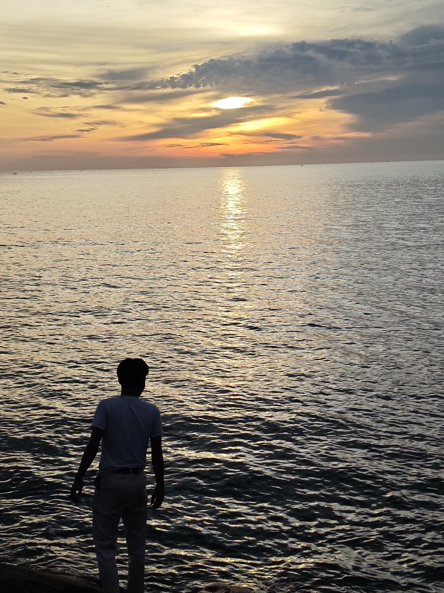The image depicts a solitary male figure standing on the edge of a body of water at sunset in Lhokseumawe, Indonesia. The individual is observed from the rear, largely silhouetted, wearing a light-colored t-shirt and light trousers. The setting sun, a bright, partially obscured orb, is positioned above the horizon line, casting intense golden and orange light across the sky and creating a distinct path of shimmering reflection on the water's surface. Layered clouds are present, displaying a gradient from warm sunset colors near the horizon to darker grays higher in the sky. The expansive body of water exhibits gentle ripples. Multiple small, dark shapes, consistent with distant boats, are discernible on the horizon.