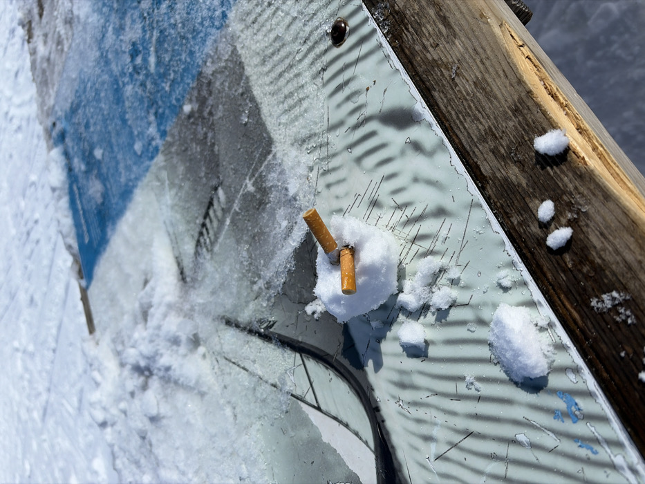 The scene depicts a small, crude snowman crafted from snow, approximately 5-7 cm in diameter, situated on an outdoor information panel within the ski resort area of La Clusaz, France. Two orange-filtered cigarette butts are embedded vertically into the snow sphere. The panel surface immediately surrounding the snow figure is a textured, pale greenish-white material exhibiting multiple fine, dark linear scratches and a single dark circular fixture to the right. This panel transitions into a surface covered by a thin layer of ice and snow, beneath which a graphic or map is discernible, featuring dark linear patterns and blue sections, indicative of a ski resort map. The background consists entirely of undisturbed white snow, typical of a ski slope or open snowy area, under bright daylight conditions. To the right, a weathered wooden plank, likely part of a bench or railing, is also partially snow-dusted. No individuals are present.