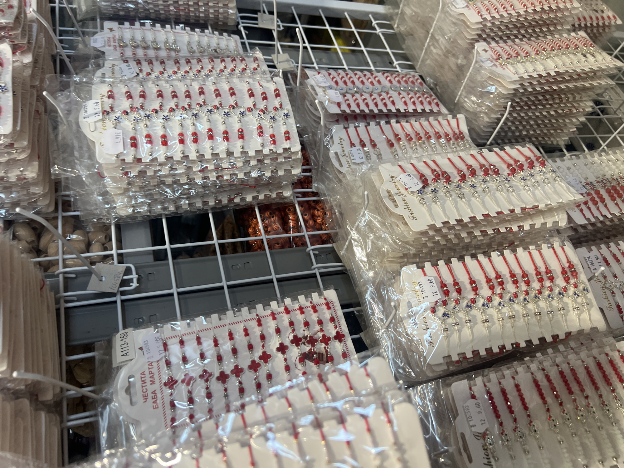 The image shows a shelf in what is likely a store in Burgas, Bulgaria, filled with stacks of "Martenița" bracelets in individual plastic bags. These bracelets, traditionally red and white, are given as gifts on March 1st and worn throughout the month. The bracelets are arranged in neat rows, with price tags visible on some of the packages. Some of the designs include beads, red crosses, and silver charms. The shelf itself is made of white wire mesh, and other packaged items can be seen in the background.