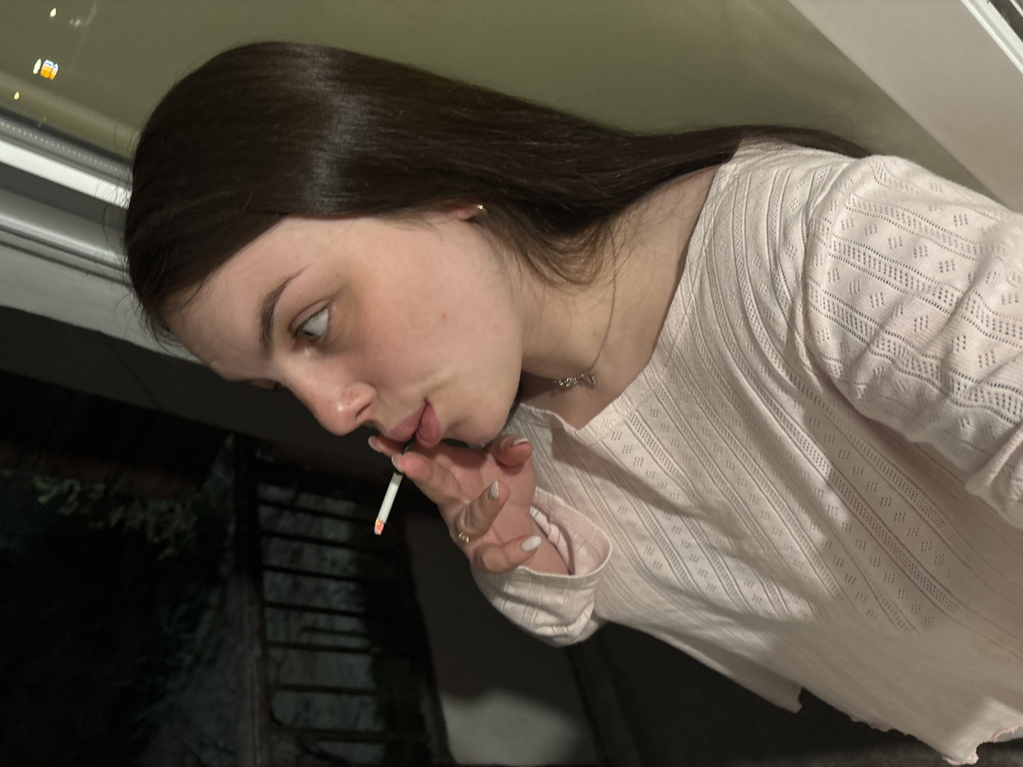 A young woman is holding a lit cigarette to her lips. She has long brown hair, light skin, and wears a light pink patterned shirt. She is wearing a necklace and ring. The background shows a window and what appears to be a dark outdoor space with some greenery. The city of Chornomorskoe, Ukraine, is not evident in the image.