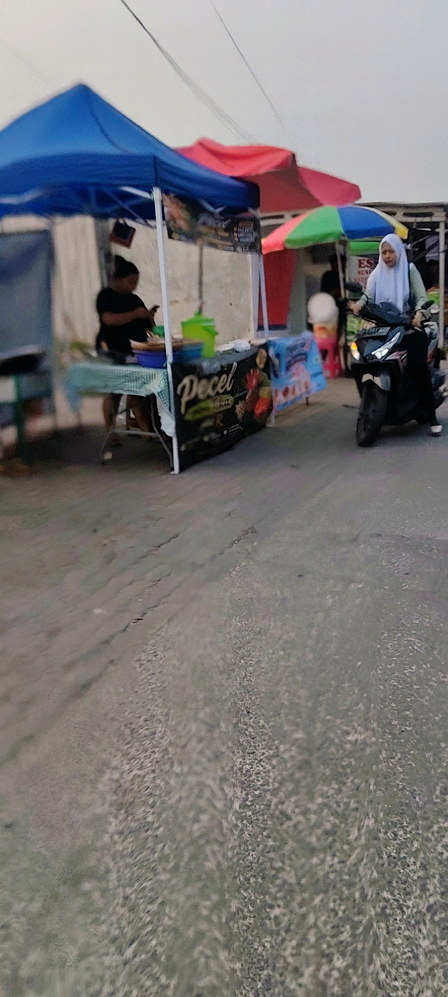 In Mekarmukti, Indonesia, a street scene unfolds with a food stall as the focal point. A person in a black shirt is seated behind a table laden with food containers and a green bucket, engaged with their phone. To the right, a woman wearing a light blue hijab is riding a black motorcycle, looking towards the left. The stall is adorned with a banner that reads "Pecel" and features images of food. Behind the stall, a red and a green and blue umbrella can be seen. The ground is paved with asphalt. The image captures a moment of everyday life at a roadside vendor.