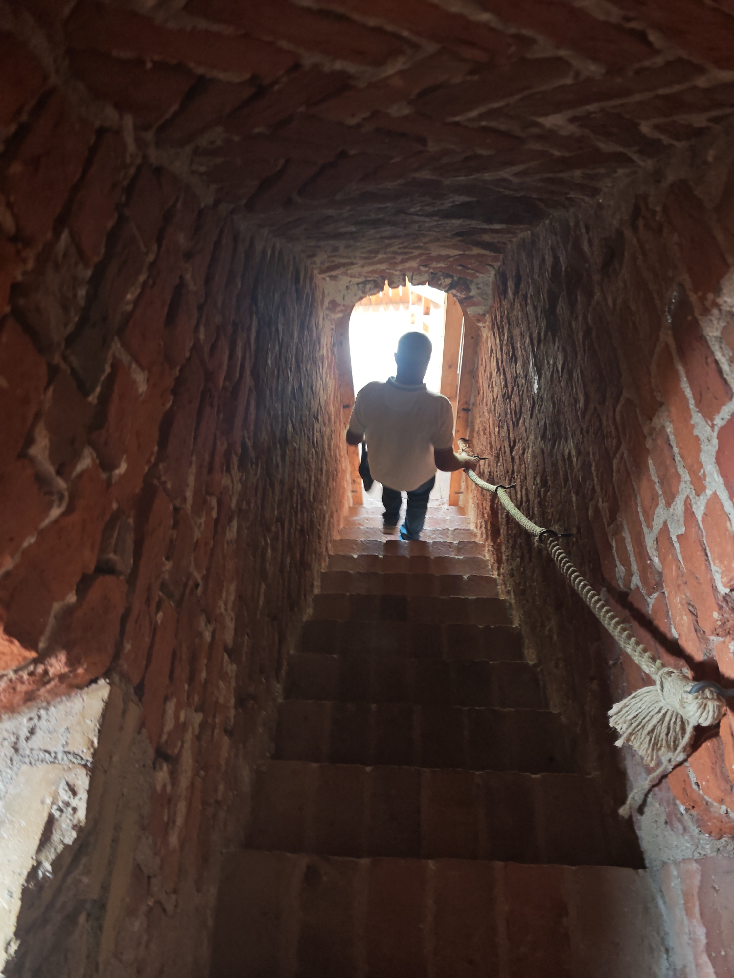 A person walks down a narrow brick staircase in a tower. The staircase is dimly lit, with natural light coming from an opening at the top of the stairs. The walls of the tower are made of red brick, and the stairs are also made of brick. A thick rope handrail runs along the right side of the staircase, secured by metal brackets. The person is wearing a light-colored t-shirt and dark pants. They are walking away from the camera, towards the light. The overall scene is one of a historic or ancient structure.