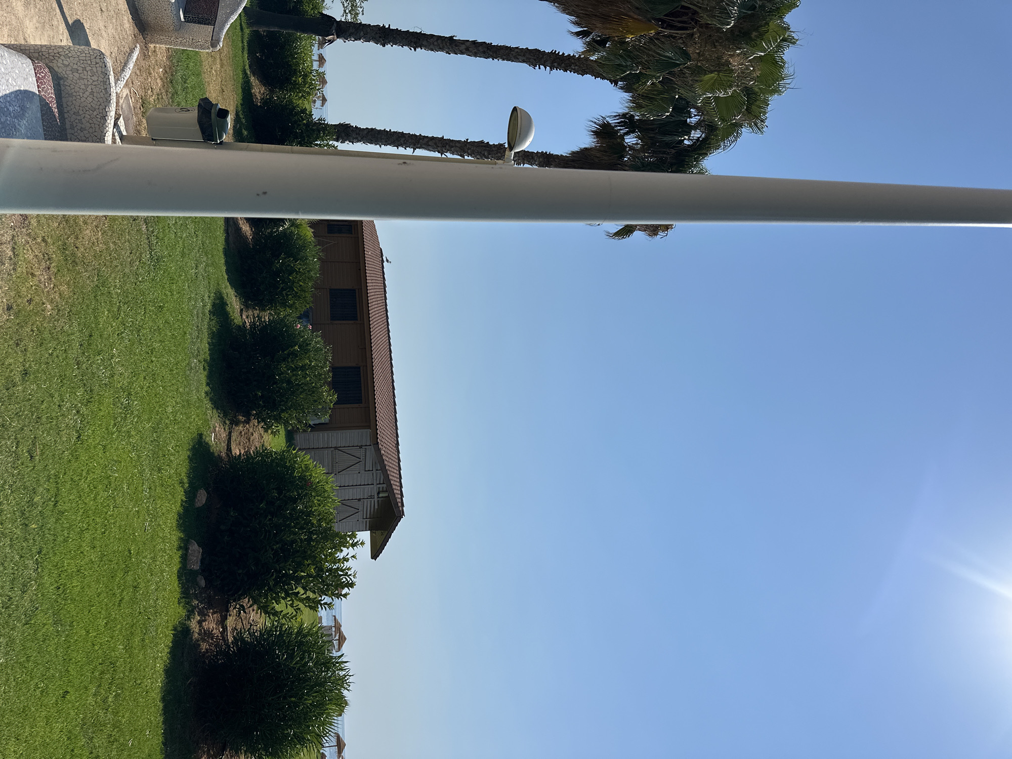 This is a landscape photograph taken outdoors in Orpesa, Spain. The image is oriented vertically, with the right side of the scene appearing at the top of the frame. The foreground is dominated by a large, white cylindrical object, possibly a pole or a structure, running horizontally across the image.
To the left of this object, there is a neatly mowed green lawn. Beyond the lawn, a cluster of three rounded, dark green bushes are visible. Further back, a rustic wooden building with a red tiled roof is situated. The building has several windows and what appear to be sliding doors or shutters with wooden slats.
In the background, the bright blue sky indicates a clear day. Two tall palm trees are visible, with their fronds reaching upwards. One palm tree is closer to the building, and the other is further to the left. A white light fixture is attached to a thin pole, positioned between the palm trees and the building, on top of the white cylindrical object.
The sun is shining brightly, casting shadows and creating lens flare in the upper right quadrant of the sky. In the far distance, beyond the bushes and the building, a glimpse of the sea and what appear to be small beach umbrellas can be seen. The overall impression is a sunny day at a seaside location, possibly a resort or a park. There is no visible text in the image. The scene conveys a sense of tranquility and a warm, pleasant atmosphere.