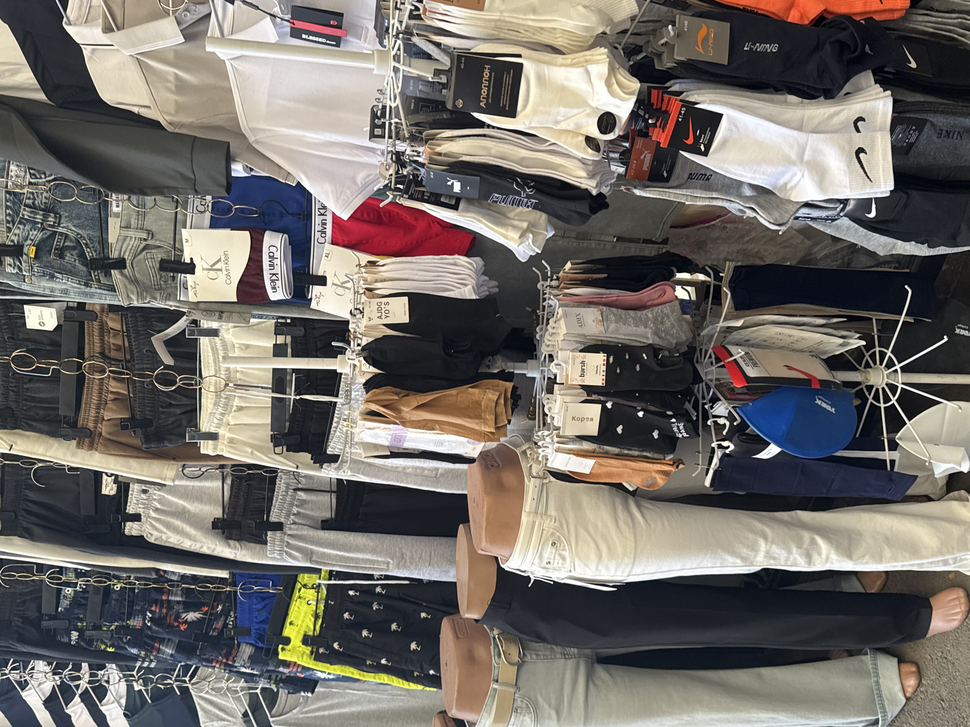 This is a full-shot, eye-level, outdoor photograph of a clothing stall in Bishkek, Kyrgyzstan. The stall is filled with various garments including pants, shorts, underwear, shirts, and socks, neatly displayed on hangers and racks. Several mannequins are visible, wearing pants and belts. The lighting suggests it is daytime. The scene appears to be a commercial area, likely a market or street vendor.
