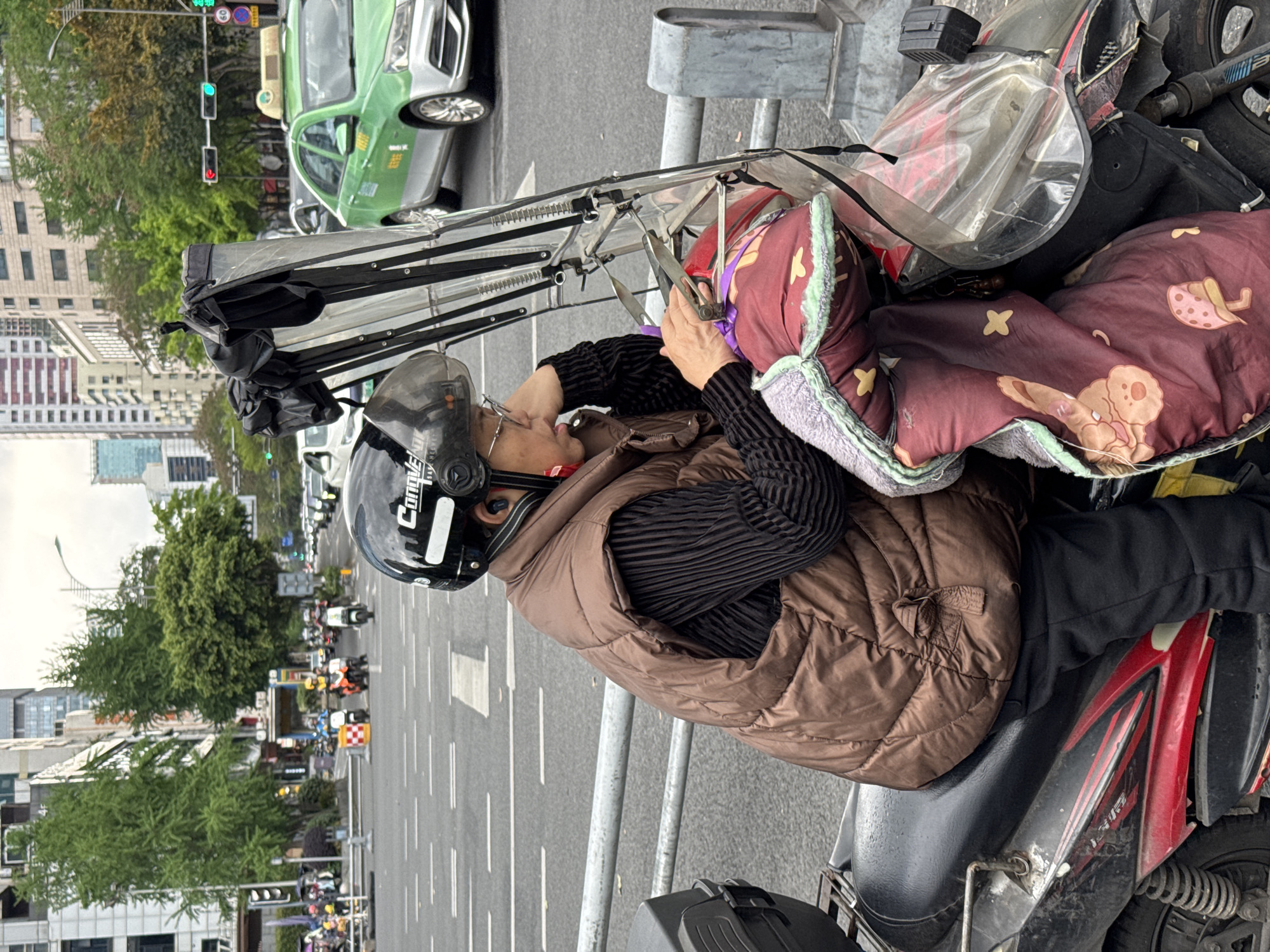 The photograph, taken in Jinjiang District, China, shows a person on a motorized scooter. The person wears a black helmet and a brown puffy vest over a black long-sleeved shirt. They also wear glasses. The scooter has a clear plastic and metal canopy attached to the front, with a brown and red patterned blanket draped over the front. The person is using a tool, possibly pliers, near the blanket and canopy. In the background, there is a busy street with cars and other scooters visible, along with trees and buildings. A green car is stopped at a traffic light in the near background. The scene suggests a street-side repair or adjustment being made to the scooter's canopy.