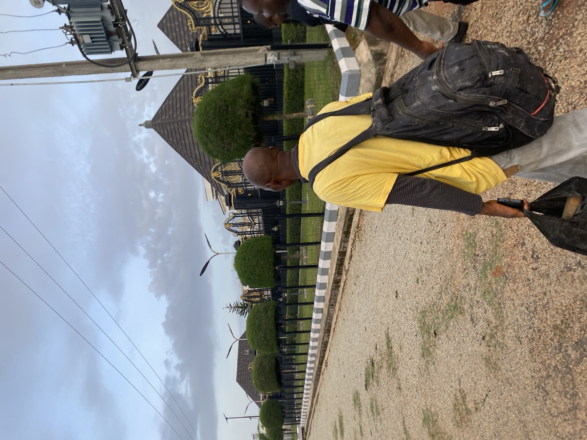 The image depicts an outdoor scene in Barikin Jirga Summa, Nigeria. A man wearing a yellow t-shirt and a black backpack walks away from the camera on a gravel path. He is carrying a black plastic bag in his left hand and a mobile phone. Another person is partially visible to his right, wearing a striped shirt. In the background, there is a black metal fence with ornate gold detailing, manicured trees, and a building with a distinctive domed roof. A utility pole with a transformer is also visible to the right of the frame. The sky is overcast.