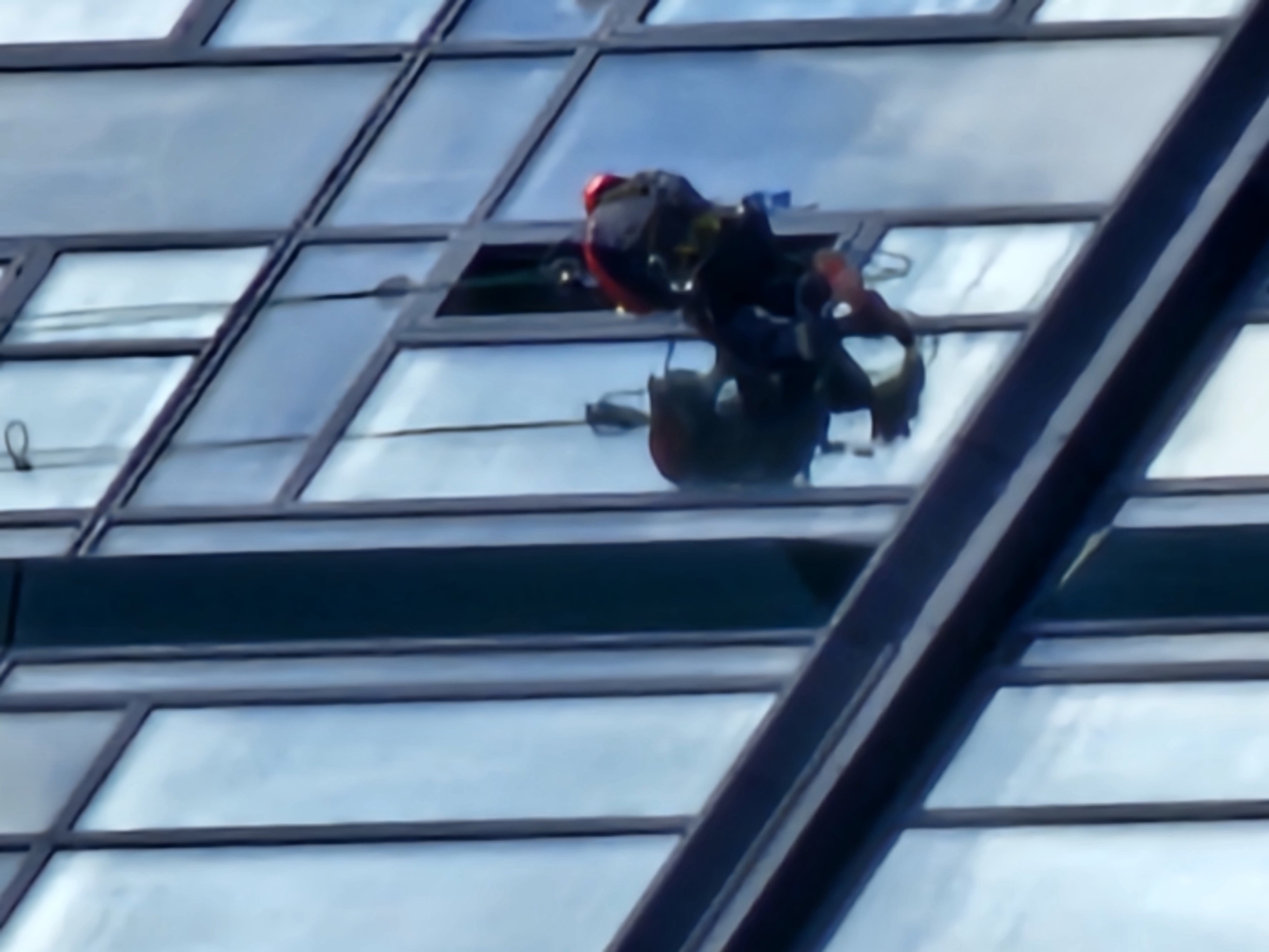 The media file shows a high-angle, medium shot of a window cleaner rappelling down the side of a modern glass-covered building. The cleaner is wearing a red helmet and dark clothing, with a harness and ropes securing them to the building. They are positioned on the side of the structure, appearing to be in the process of cleaning or inspecting the windows.
The building's facade is composed of many rectangular glass panes framed by dark-colored mullions, suggesting a contemporary architectural style. The reflections in the glass indicate an overcast sky, with soft, diffused light, implying it is daytime, possibly mid-morning or afternoon. The setting appears to be an urban environment, given the scale of the building and the presence of such a structure in Moscow, Russia.
The overall impression is one of a routine, albeit high-risk, professional activity taking place in an urban setting. There are no visible signs or text within the frame. The image captures a moment of work at height, emphasizing the precarious nature of the task.