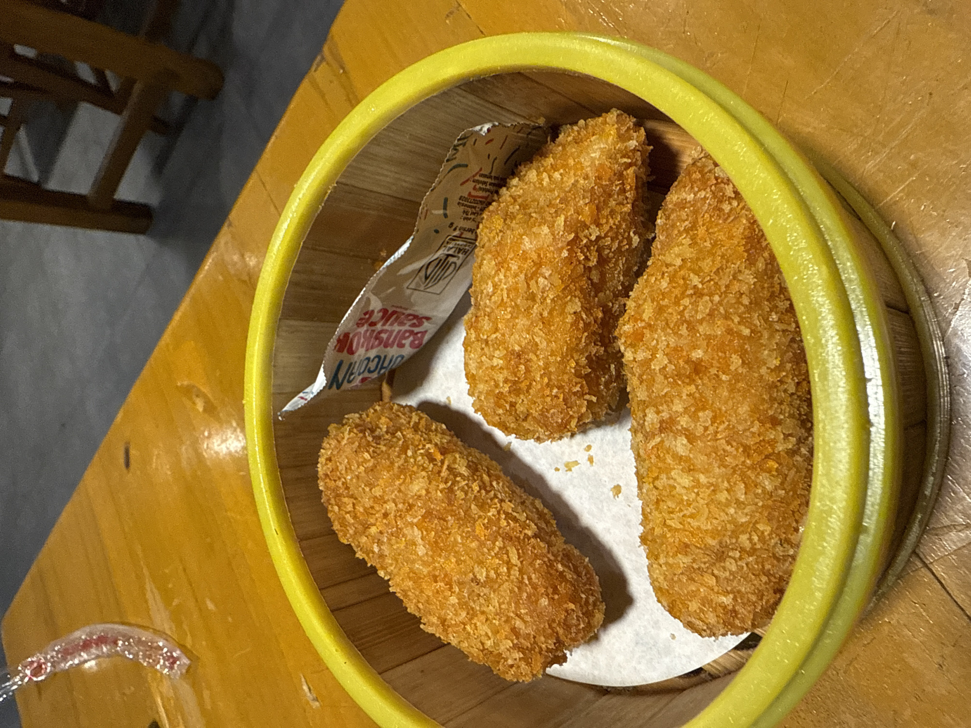 A close-up shot captures a bamboo steamer basket filled with three golden-brown, breaded items. These items appear to be fried snacks, possibly croquettes or spring rolls, arranged on a white paper liner within the steamer. A small packet of "Bangkok Sauce" is partially visible, peeking out from under one of the fried items. The steamer basket itself has a bright yellow rim. The entire basket rests on a warm-toned wooden table. In the background, a portion of a wooden chair or table leg is visible, suggesting a dining setting. The lighting is warm and indoor, casting soft shadows and highlighting the crispy texture of the food. The overall impression is one of a casual dining experience, likely a meal or appetizer. The visible text on the sauce packet includes "Bangkok Sauce" and what appears to be a halal certification logo.