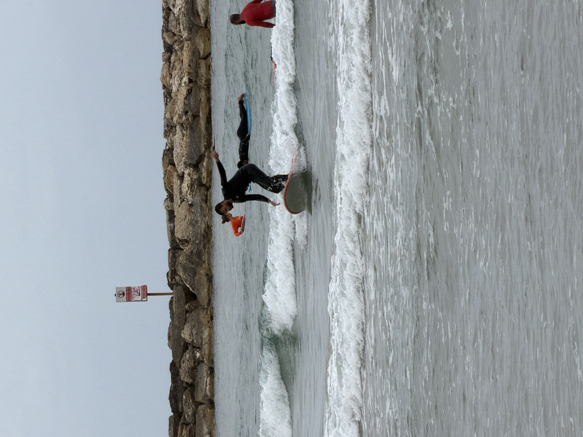The image shows a man in a black wetsuit surfing on a bodyboard in the ocean. He is positioned near a stone wall that appears to be a breakwater or jetty. To the left of the wall, a sign is visible. Further into the water, another person on a boogie board and a person wearing a red shirt can be seen. The scene likely takes place in Tel-Aviv, Israel, as it's a popular coastal city known for its beaches. The environment consists of the ocean, waves, and the stone breakwater. The primary action is the man surfing on the bodyboard.