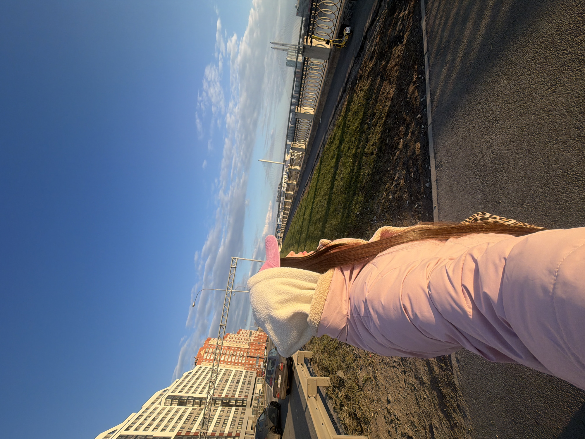 The image shows a person wearing a pink coat and a white hooded jacket with furry trim, standing on a sidewalk or path. The perspective is canted, with the sky and buildings tilted. The sky is predominantly blue with some clouds. In the background, there is a building with a multi-story structure and light-colored facade, as well as a street with parked cars. In the distance, there is a bridge spanning a body of water, with the skyline of Saint Petersburg, Russia, visible. Along the path, there is a metal railing and a strip of grass. The lighting suggests it is daytime.