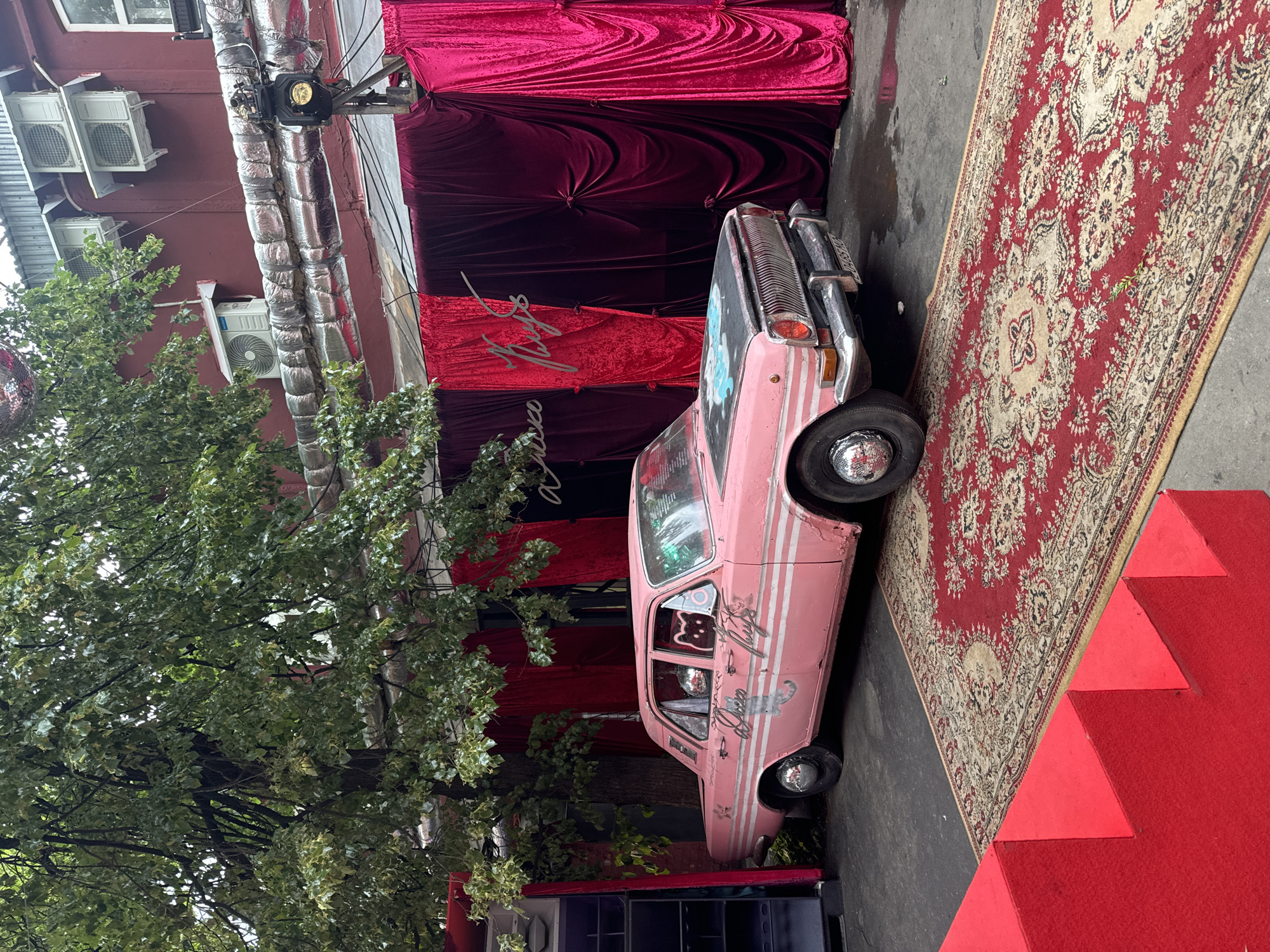 The image depicts an outdoor scene in Moscow, Russia, likely a venue or event space, characterized by a vibrant and eclectic setup. In the foreground, a bright red staircase leads up to a grey asphalt surface. A large, ornate red carpet with intricate beige and black patterns is laid out on the asphalt, extending towards the background.
Dominating the central part of the scene is a vintage pink car, which appears to be a Gaz-24 Volga. The car is adorned with custom decals, including white stripes along its side and a cartoonish cat illustration with a disco ball inside its head on the rear passenger window. The hood of the car features a painted design, possibly a light blue cloud-like image. The car's chrome hubcaps and grille are visible.
Behind the car, a dramatic backdrop of rich velvet curtains in shades of deep red and fuchsia is displayed. The curtains are draped and tied back, creating a theatrical effect. A metallic sign with stylized lettering is mounted on the curtains, appearing to read "Disco Club" or a similar phrase in Russian.
To the left of the car, a tall, dark structure resembling a DJ booth or speaker system is partially visible, with blue lighting effects. Above the car and curtains, the exterior of a building is seen, painted in a dark red hue. Several air conditioning units are mounted on the wall, and large, insulated pipes run horizontally across the facade. A spotlight is positioned on a metal stand, directed towards the area.
A large, leafy tree with abundant green foliage arches over the scene from the left, providing shade. The ground appears slightly wet, suggesting recent rain. The overall impression is one of a unique and visually striking outdoor entertainment or event setting.