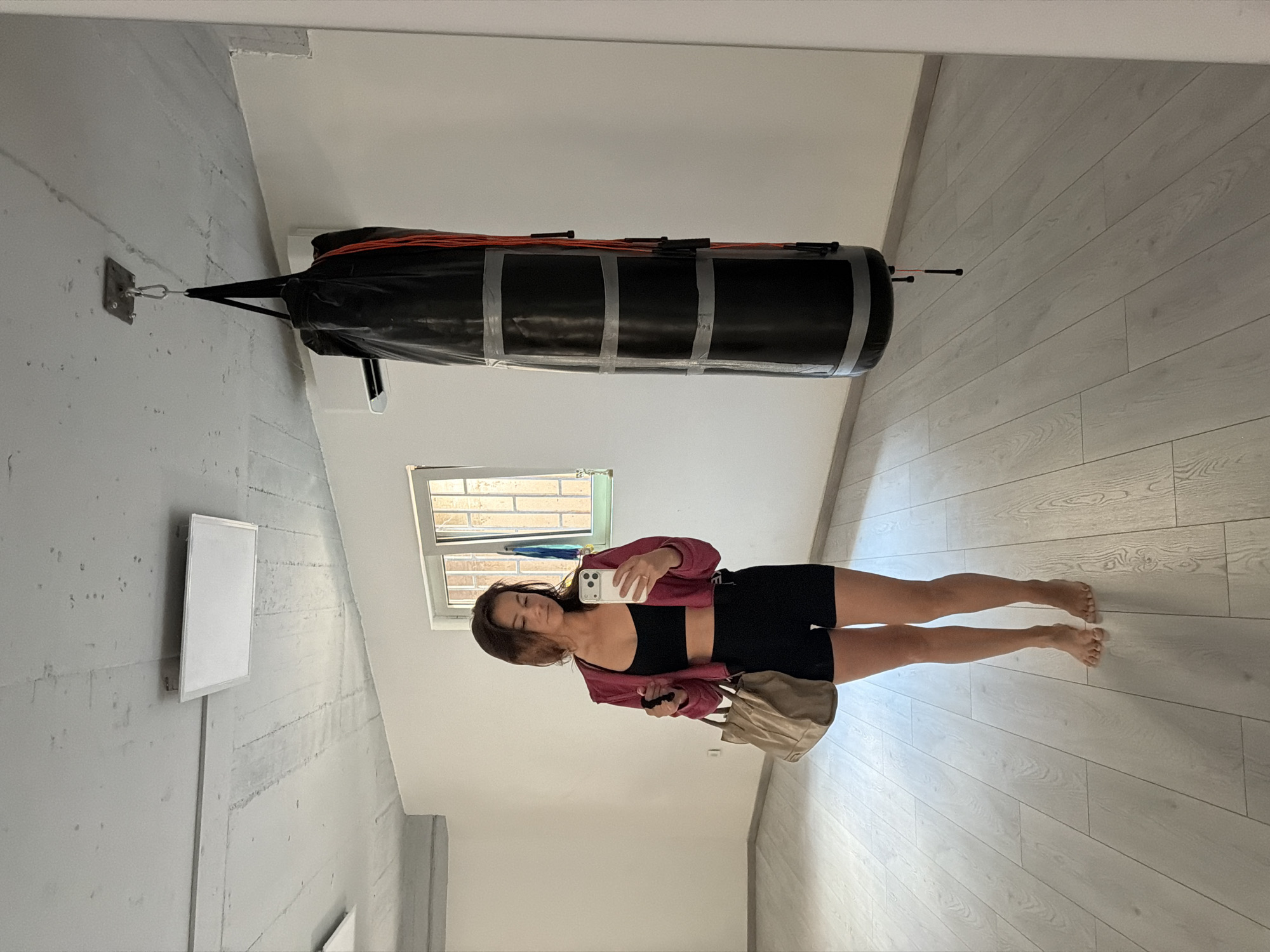 A young woman takes a selfie in a room with a punching bag suspended from the ceiling. She stands barefoot on a light wood floor, wearing a black sports bra, black shorts, and a burgundy jacket. She holds a white phone in her left hand and a beige bag in her right. The punching bag is black with silver duct tape around it. The room has white walls and a window with a view of an exterior, and the setting appears to be an indoor workout space or a room being used for fitness. While this is an indoor scene, there is not enough information to confirm a location in Budva, Montenegro.