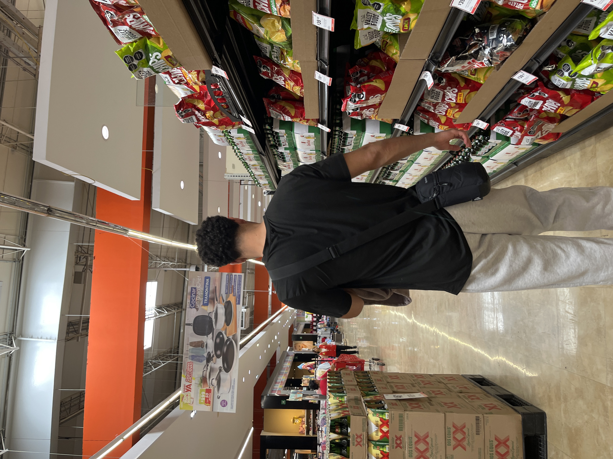 The image shows an individual shopping in a grocery store. The person is wearing a black t-shirt, light gray sweatpants, and has a black bag slung across their body. They are standing in front of a shelf stocked with various brands of chips, including Valley Foods and others labeled in Spanish. The price tags on the shelf indicate costs of $39.00 and $39.90. The shelves above also hold similar products.
Further down the aisle, other shoppers can be seen, along with displays of merchandise like cookware from the brand Tramontina. Stacks of Dos Equis beer are also visible in the lower part of the frame. The store has bright lighting with visible ceiling structures and orange accents on the walls.