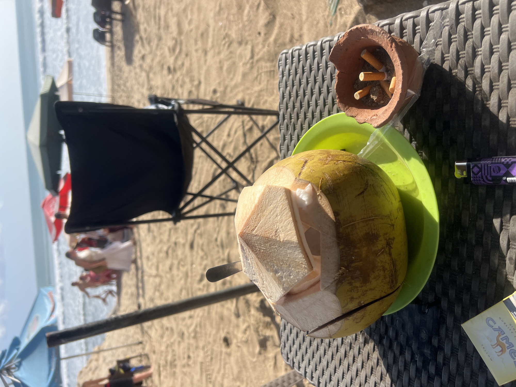 A close-up shot of a partially opened coconut with a metal straw inserted into it, placed on a green plate. To the right, a small, rustic ashtray made of clay holds several cigarette butts. A purple and black lighter rests on the patterned table in the foreground, next to a yellow cigarette pack labeled "CAMEL." In the background, the beach of Kuta, Indonesia, is visible, with umbrellas, people walking by the water, and the ocean with gentle waves. The lighting suggests it is a sunny day.