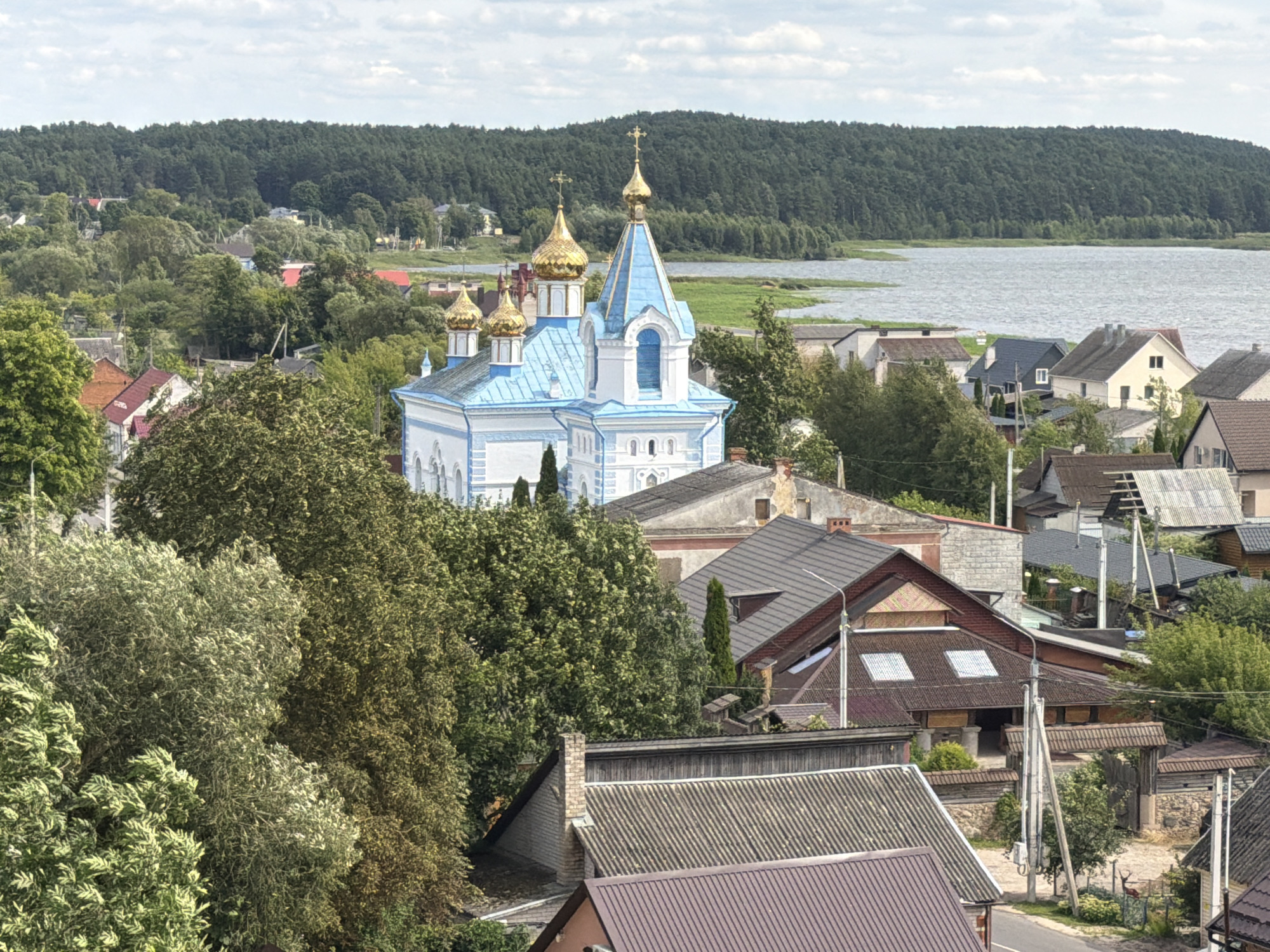 A high-angle, eye-level, medium shot captures a picturesque view of a village nestled beside a tranquil body of water, with a dense forest forming a verdant backdrop under a cloudy sky. The focal point of the scene is a charming blue and white church adorned with golden onion domes, its intricate architectural details standing out against the natural landscape.
Surrounding the church, a cluster of houses with red and brown tiled roofs are visible, interspersed with lush green trees that create a sense of depth and tranquility. The houses are of varying sizes and architectural styles, suggesting a well-established community.
In the foreground, the rooftops of several houses are prominent, some featuring weathered grey tiles and others with more modern brown coverings. The scene is characterized by its peaceful ambiance, with no people or animals visible, allowing the viewer to appreciate the serene beauty of the village and its natural surroundings. The overall impression is one of a quiet, idyllic rural setting, with the church serving as a spiritual and architectural anchor.