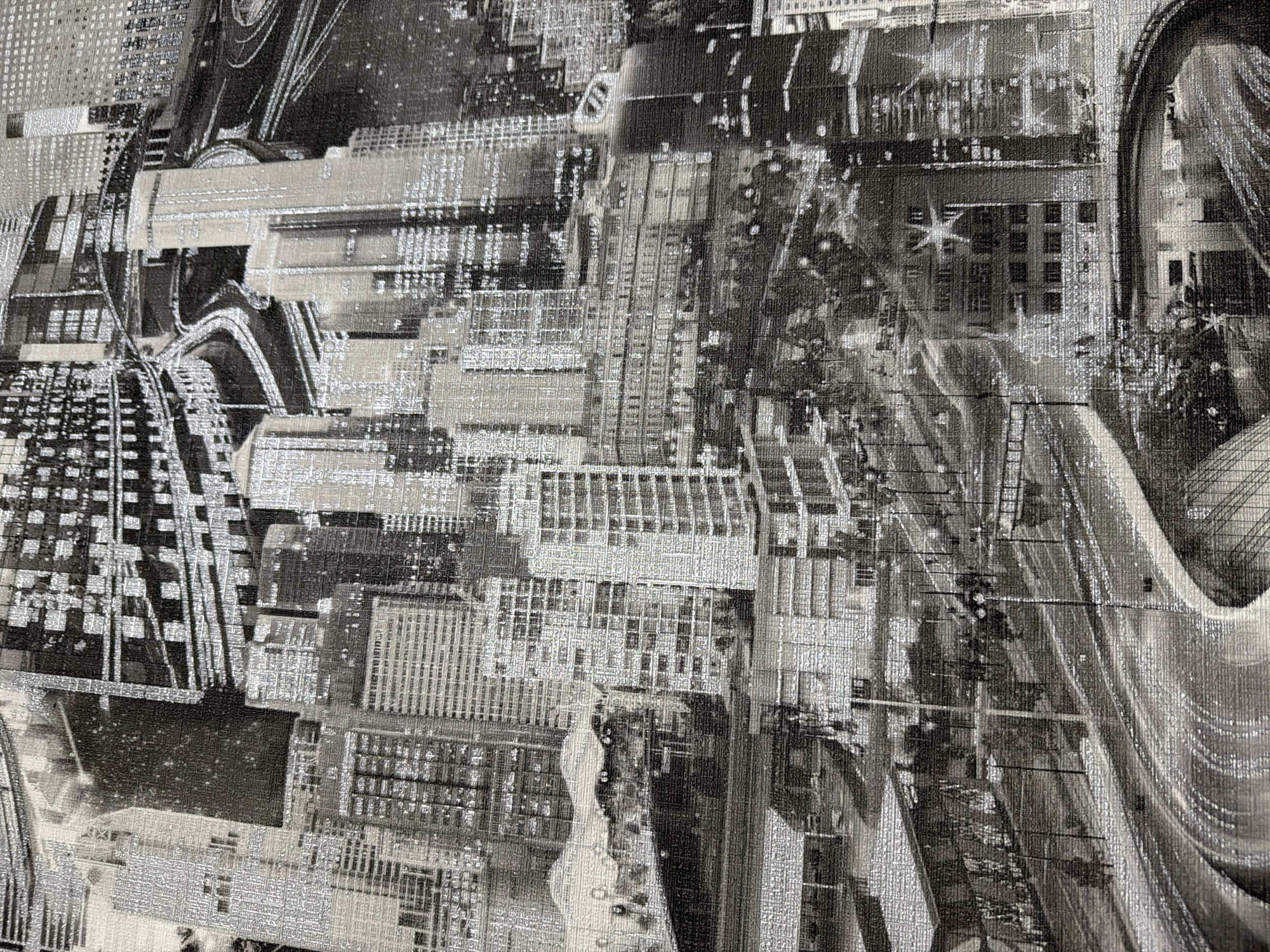 The image displays a close-up, high-angle view of a city skyline rendered in a grayscale, textured fabric. The design features numerous tall buildings with detailed window patterns, suggesting a dense urban environment. Intersecting roadways, likely elevated highways, are prominent, weaving through the structures. The overall impression is one of a bustling, modern metropolis, possibly at night, given the shimmering, reflective quality of the fabric which might represent lights. There are no people visible in the image. The scene is purely an artistic depiction and not a photograph of a specific location in Mali Kosharyshcha, Ukraine, or any other real place.