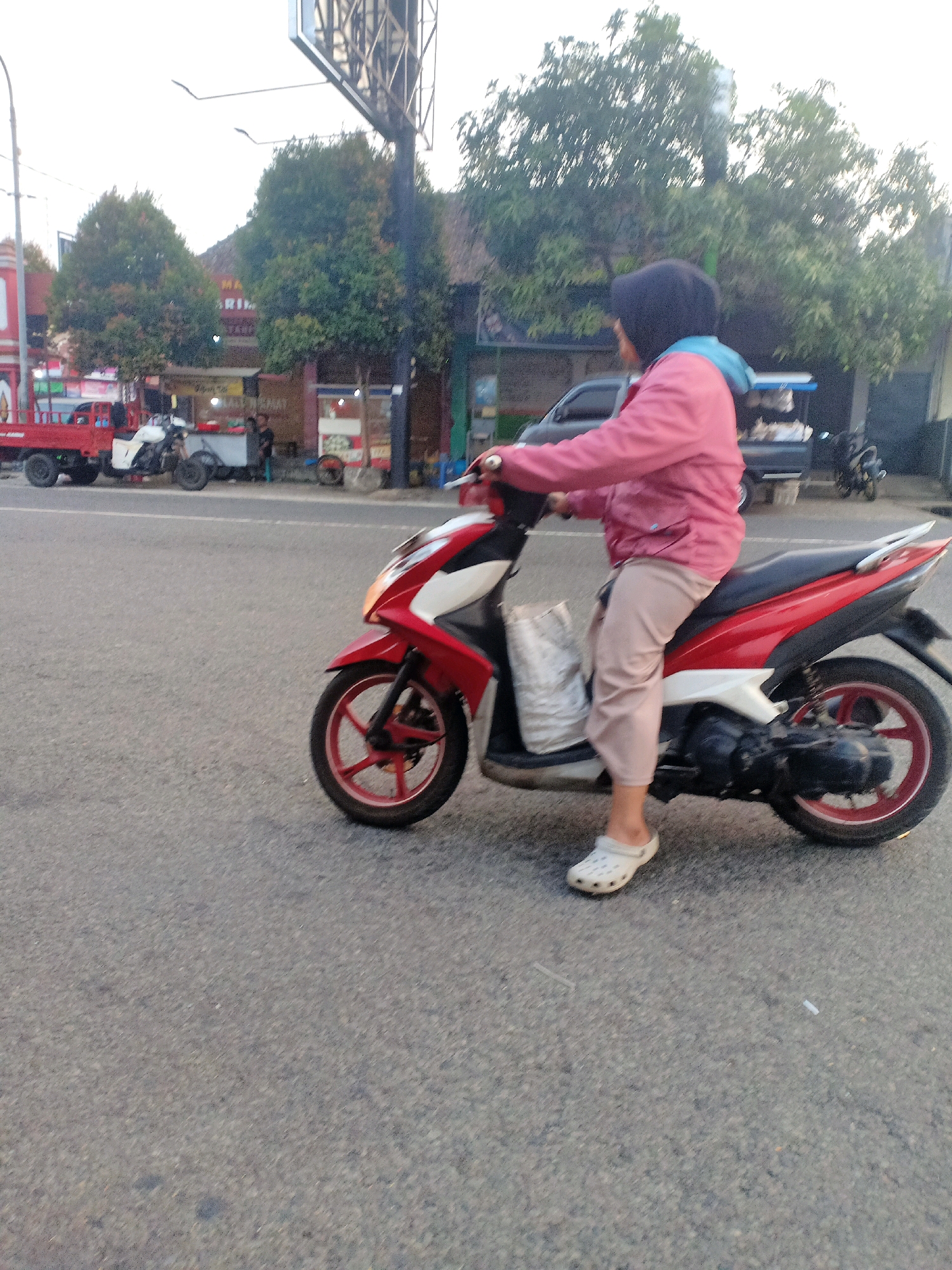 In Mojoreno, Indonesia, a woman wearing a pink jacket, light-colored pants, and white slip-on shoes rides a red and white scooter on a paved road. The woman has a dark hijab covering her hair. A white bag is secured to the front of the scooter. In the background, a red truck is parked on the side of the road next to a white motorbike. Beyond the vehicles, there are several trees and buildings, including a structure with a sign that appears to have Indonesian text. The sky is bright, suggesting it is daytime.