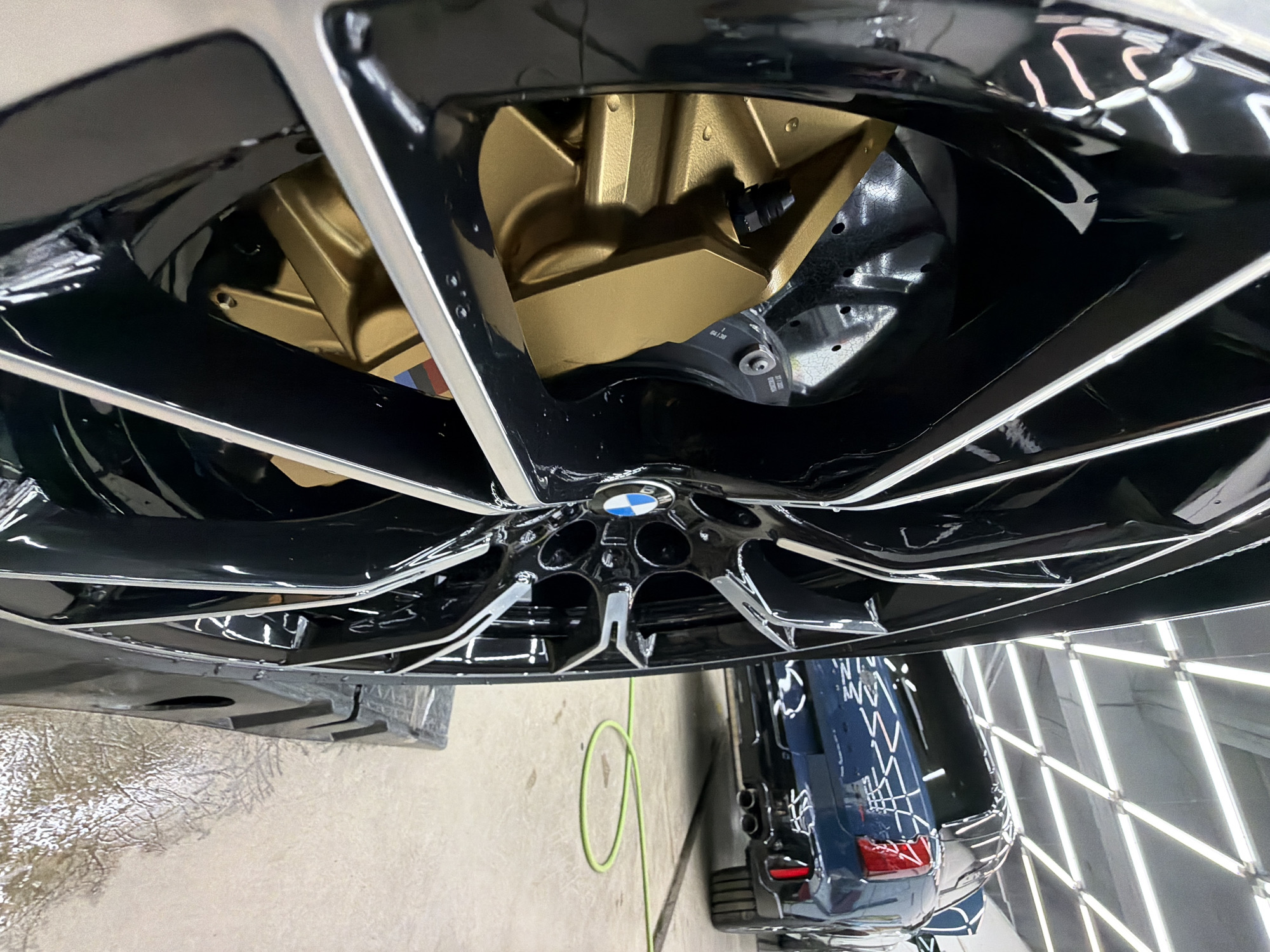 The image shows a close-up of a black BMW wheel with gold brake calipers. In the background, a dark blue SUV is visible. The scene appears to be in a well-lit garage or workshop, with fluorescent lights on the ceiling. A green hose is lying on the concrete floor. The image is taken from a low angle, emphasizing the wheel and brake system. There are no people visible in the image. The location of Krasnoyarsk, Russia, is not discernible from the visual information provided in the image.
