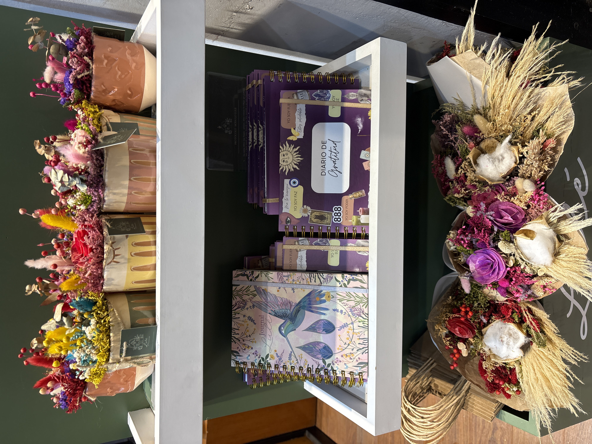 On a white shelf in a store, three decorative pots filled with colorful dried flowers are displayed. The pots are made of ceramic with a textured finish and feature various patterns. The flowers are a mix of bright reds, yellows, blues, purples, and pinks, creating a vibrant and whimsical arrangement.
Below the pots, on another white shelf, are several spiral-bound notebooks. The notebook in the foreground has a cover with a watercolor illustration of a hummingbird and floral motifs, with the word "Journal" prominently displayed. To the right, a stack of purple notebooks is visible, some with gold trim and eye-catching designs featuring celestial bodies, tarot cards, and inspirational text in Spanish, such as "DIARIO DE Gratitud" (Gratitude Diary).
At the bottom of the image, two bouquets of dried flowers and grasses are arranged in a dark green display. These bouquets feature a rustic aesthetic with elements like pampas grass, dried roses, and cotton-like accents. The scene suggests a retail environment, possibly a gift shop or florist, located in San Isidro, Peru, a district known for its upscale boutiques and commercial establishments. The overall atmosphere is one of curated beauty and artisanal products.