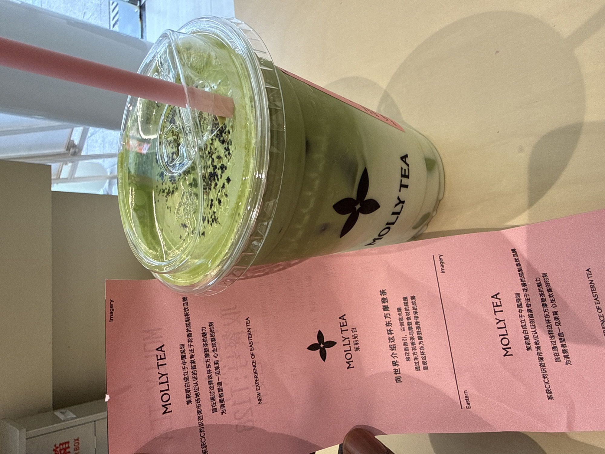 A close-up shot shows a cup of matcha tea from "Molly Tea," a tea brand founded in Shenzhen, China, resting on a pink promotional flyer or placemat with the brand's logo. The cup, which is clear plastic with a domed lid, contains a green matcha beverage with black seeds sprinkled on top. A pink straw protrudes from the lid. The flyer contains Chinese text, part of which is translated as saying the brand is "the first brand specializing in floral aroma-flavored tea drinks in the market certified by CIC insight." The background is a plain, light-colored surface, possibly a table. The image is taken indoors with ample lighting, but the angle and cropping suggest it was casually captured, potentially by a customer.