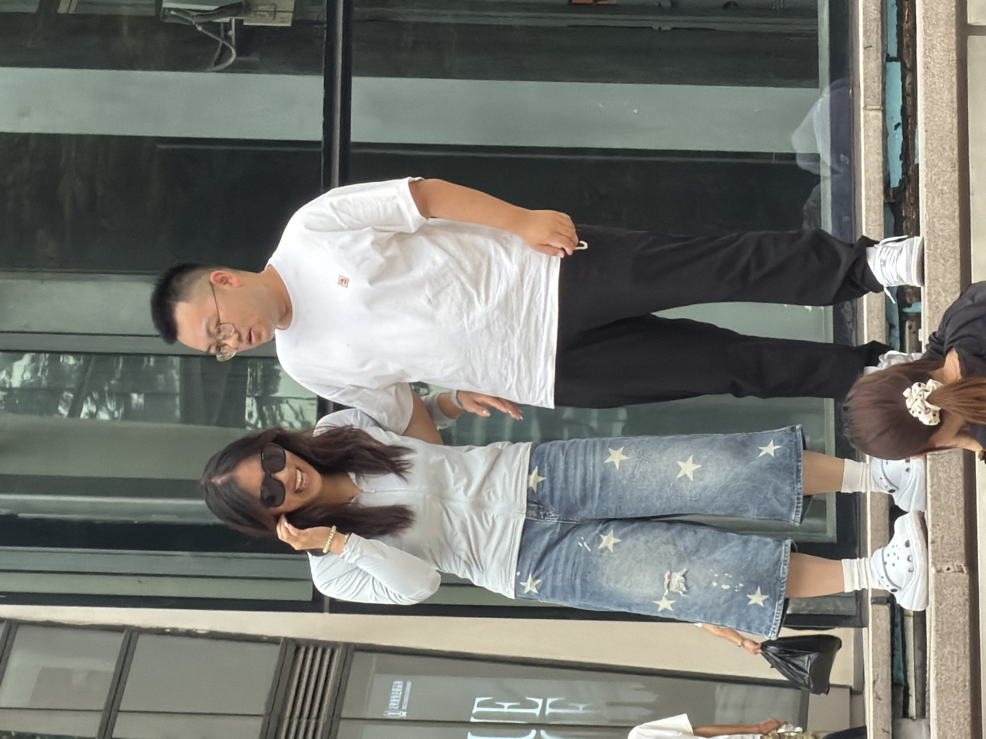 In Yuzhong District, China, a man and a woman stand on a raised platform in front of a modern building with large glass windows. The woman, on the left, wears a light blue long-sleeved zip-up top and distressed denim culottes adorned with white stars. She has on white Croc-style shoes and white socks. Large black sunglasses obscure her eyes, and she is smiling with her left hand touching her hair. A light brown bracelet is visible on her left wrist. She holds a black bag in her right hand. The man, on the right, wears a white t-shirt with a small orange logo on the left chest and black sweatpants. He has on white sneakers and appears to be looking to his left. He wears glasses. The background shows the reflective surface of the building, suggesting an urban environment.