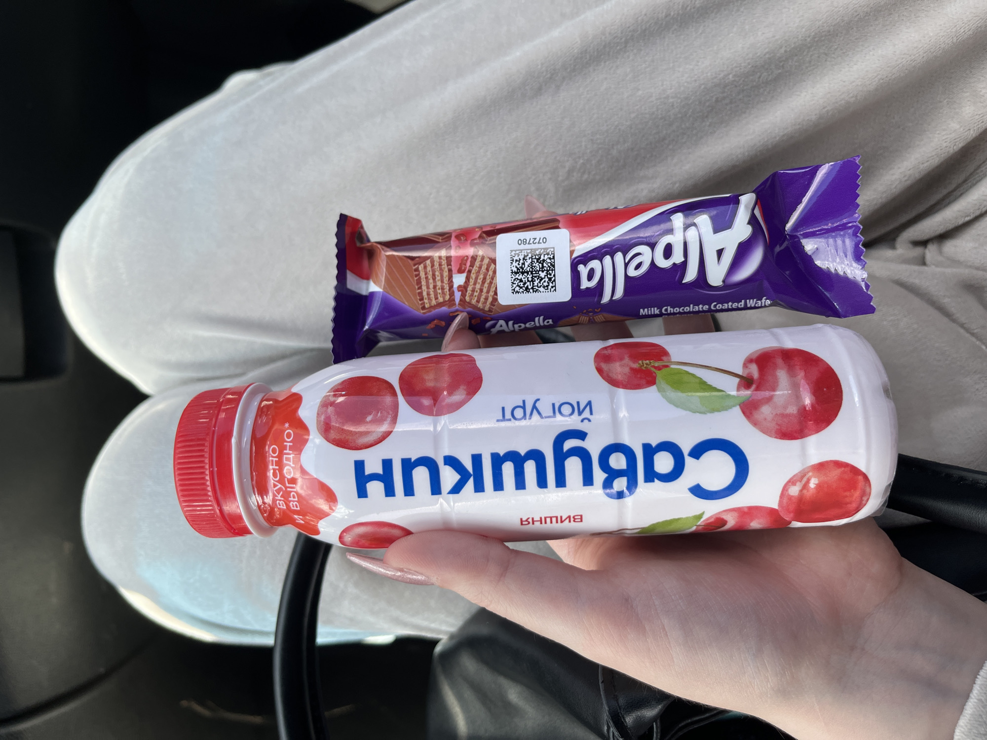 A close-up shot shows a person's hand holding a chocolate bar and a bottle of cherry-flavored yogurt. The chocolate bar, a "Alpella Milk Chocolate Coated Wafer," is purple and red with an image of the wafer inside. The yogurt bottle is white with a red cap and features an illustration of cherries. The text on the yogurt bottle is in Russian and reads "вкусно и выгодно" which translates to "tasty and profitable," and "черный" which translates to "black" or "cherry." The scene appears to be indoors, possibly in a car, given the dark background and the texture of the clothing, which looks like white sweatpants. The lighting suggests it is daytime. The person's fingernails are painted a shimmery pink. The overall impression is a casual moment of someone holding snacks.
