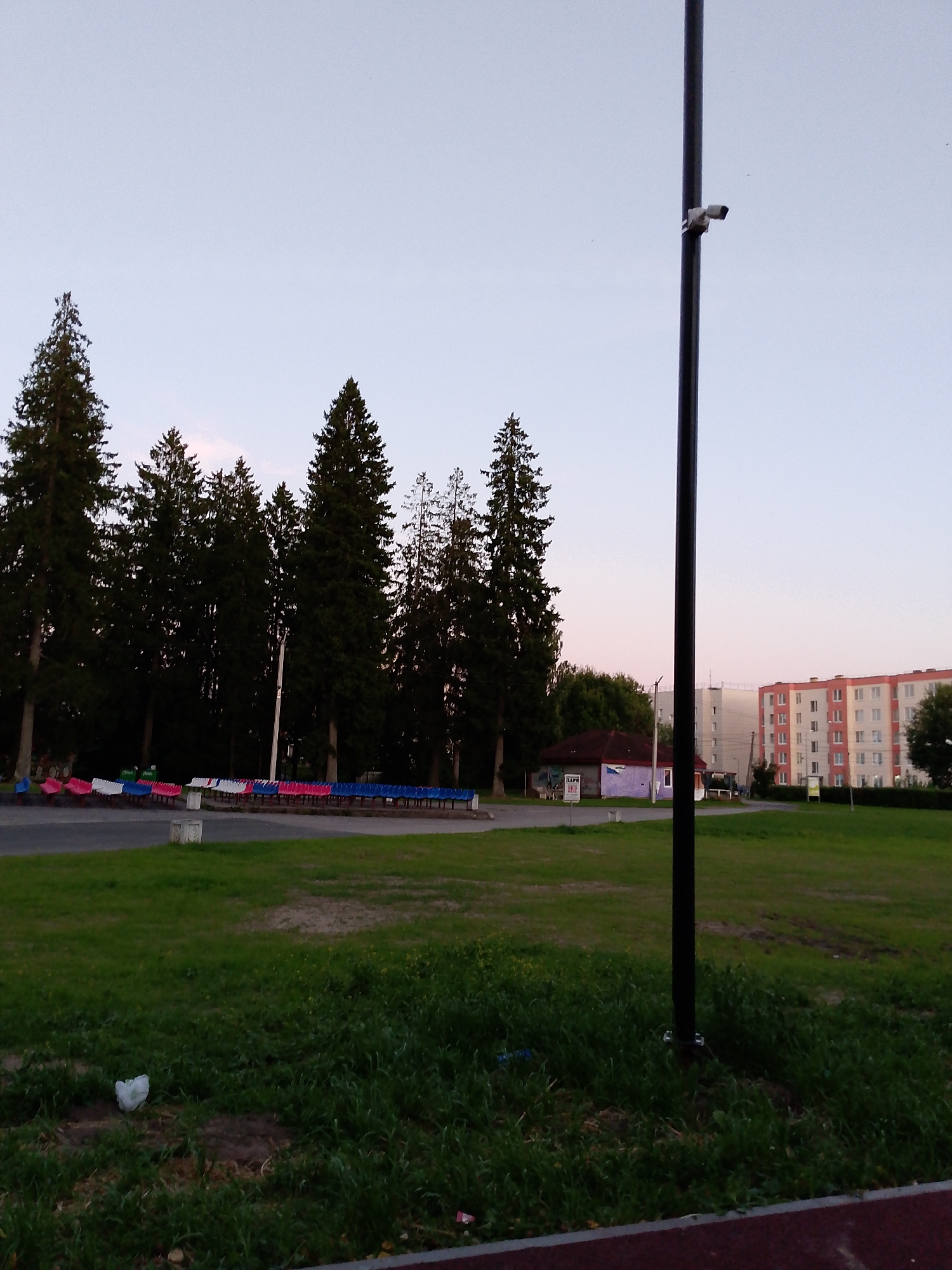 In the city of Volosovo, Russia, a street light with an attached security camera stands tall in the foreground. Behind it, a row of bleachers with red, white, and blue seats sits on a paved surface, bordering a grassy field. A small, single-story building with a red roof and purple-painted walls is visible behind the bleachers, featuring a sign that reads "ПАРК". Further in the background, two multi-story apartment buildings with cream and orange facades stand against a pale sky. Several tall evergreen trees are scattered throughout the scene, with a dense forest line to the left. The overall environment appears to be an outdoor public space, possibly a park or recreation area, as indicated by the presence of bleachers and the "ПАРК" sign.