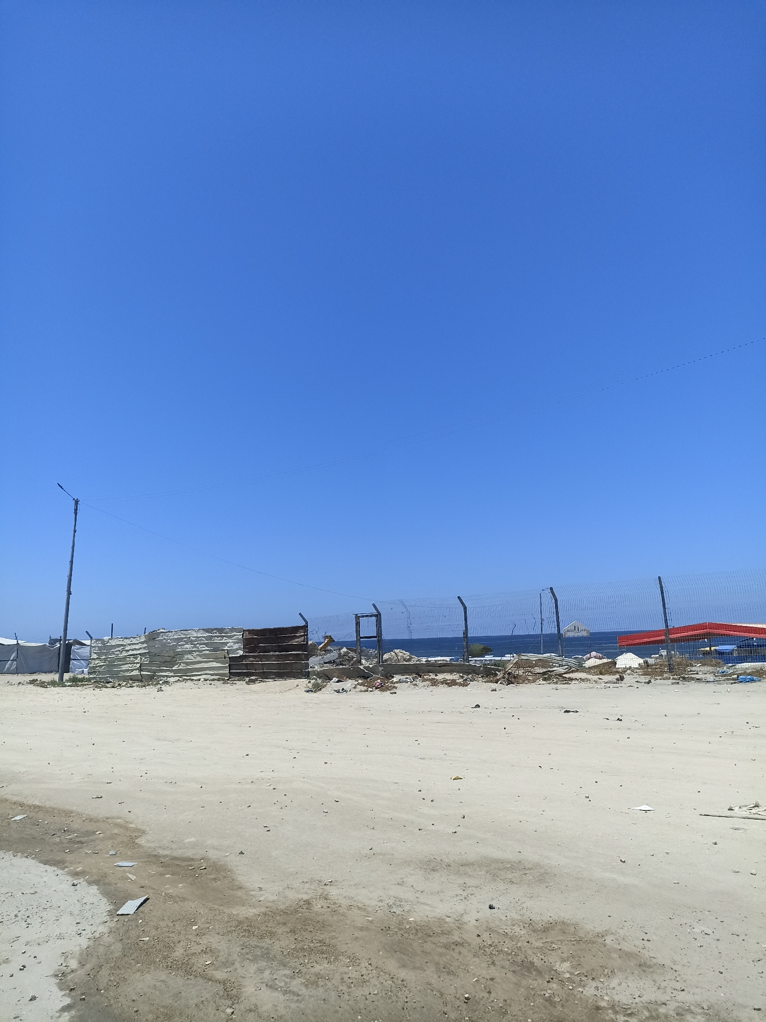 This media file depicts an outdoor scene under a clear blue sky. The foreground is a sandy, uneven ground with some scattered debris and puddles. A makeshift fence or barrier made of corrugated metal and wood runs horizontally across the mid-ground, separating the sandy area from what appears to be a body of water in the background. Beyond the fence, a large expanse of blue water is visible, with a small island or rock formation in the distance. A wire fence with metal posts also runs along the water's edge, and there are some structures or tents behind the corrugated metal barrier, some appearing to be made of white canvas. A tall light pole is situated to the left of the frame. The overall impression is one of a somewhat desolate or underdeveloped coastal area, possibly undergoing construction or in a state of disrepair. The clear sky and bright sunlight suggest it is daytime, likely a warm and sunny day. There is no visible text in the image.