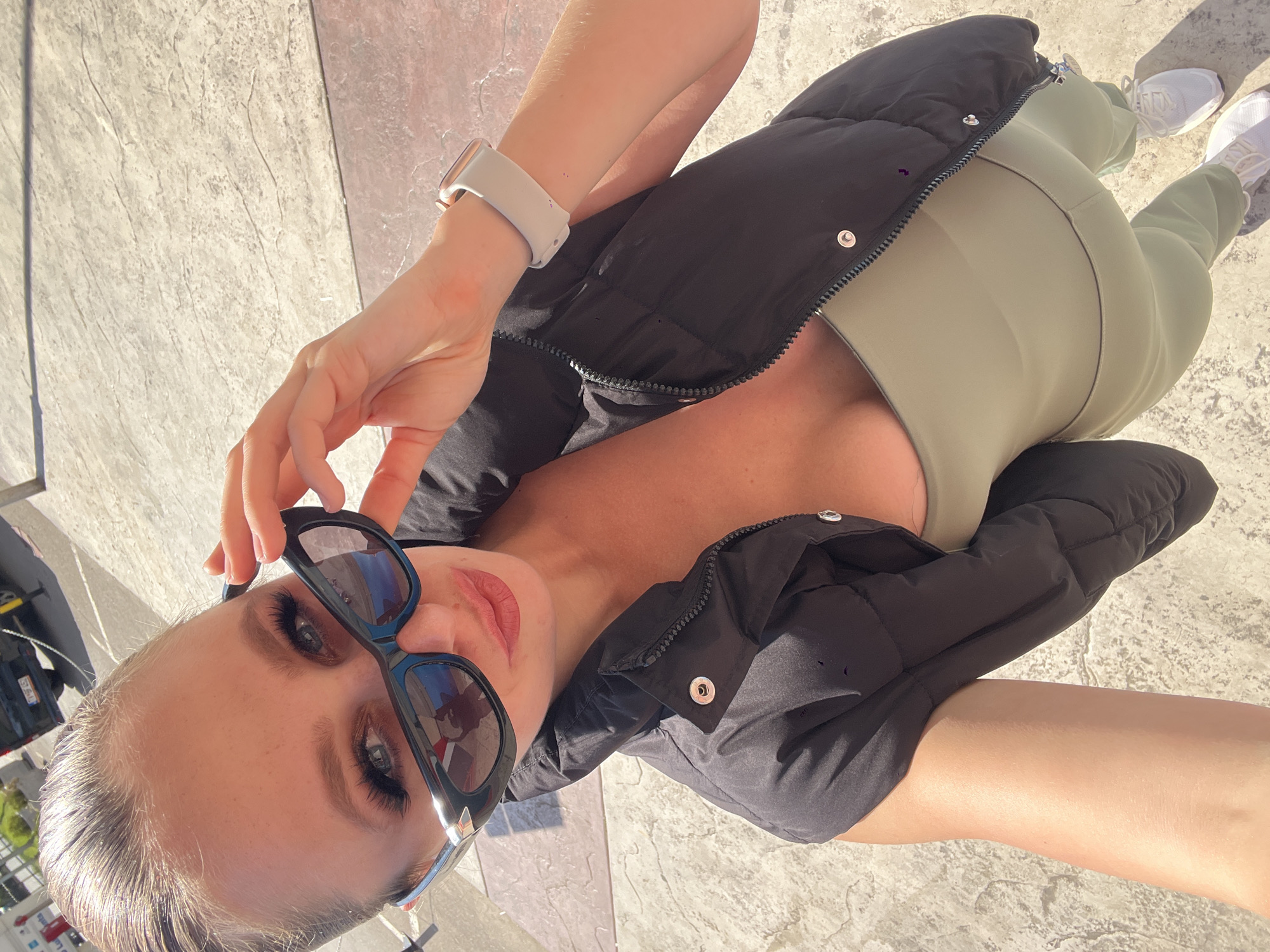 The image captures a woman from a high-angle perspective, seemingly a selfie. She is wearing a black puffer jacket over a light green top and matching leggings. Her footwear consists of white sneakers. The woman is holding a pair of sunglasses, partially obscuring her face. She has fair skin and light-colored hair pulled back. Her left wrist sports a white smartwatch.
The setting appears to be an outdoor paved area with light-colored stone tiles. There are some visible elements in the background, including a car and a building with advertisements. The general aesthetic and presence of the building with advertisements suggest the location could be in the city of Budva, Montenegro.